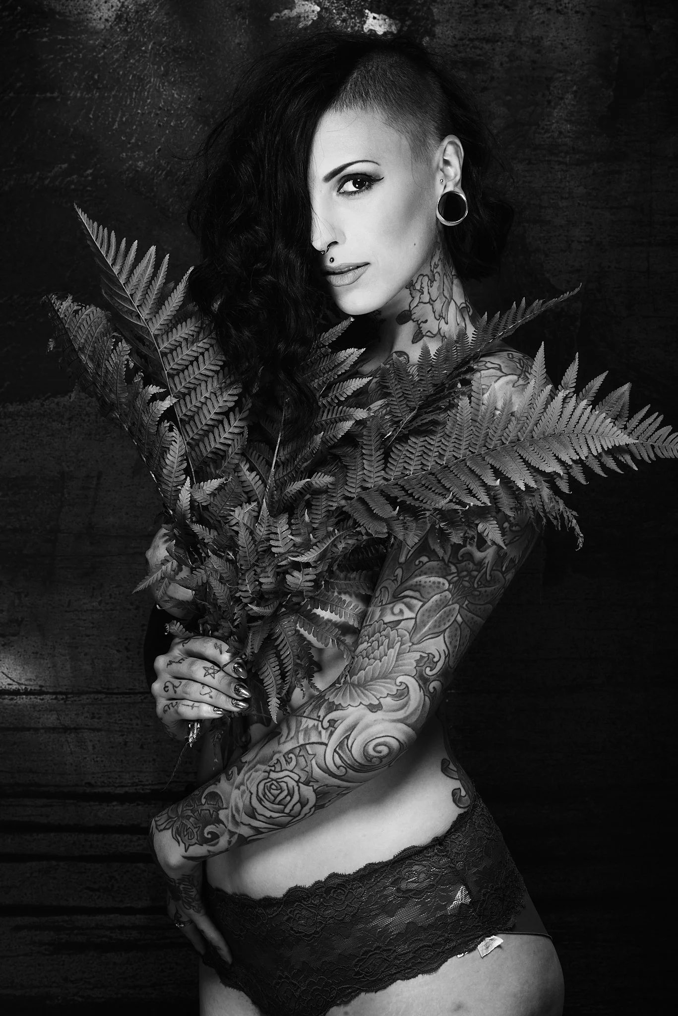 A tattooed woman with dark, wavy hair, partially shaved on one side, holding a fern plant in front of her chest, wearing lace underwear, against a dark background.