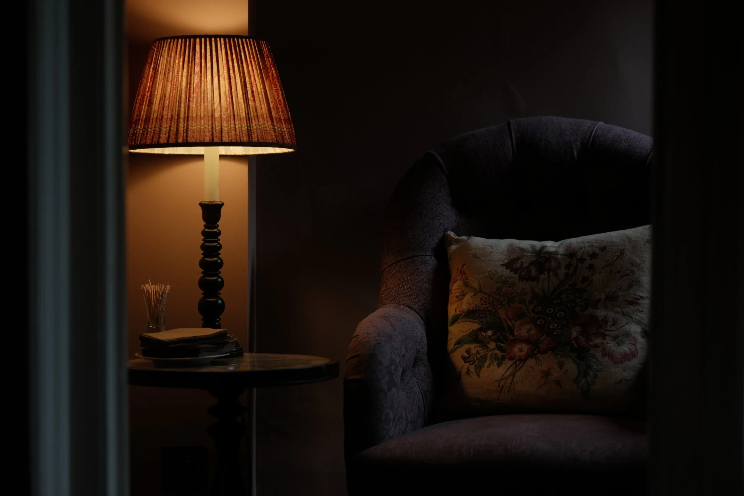 Dimly lit interior scene with a table lamp, upholstered armchair with a floral pillow, and a side table holding some papers and a small glass container.