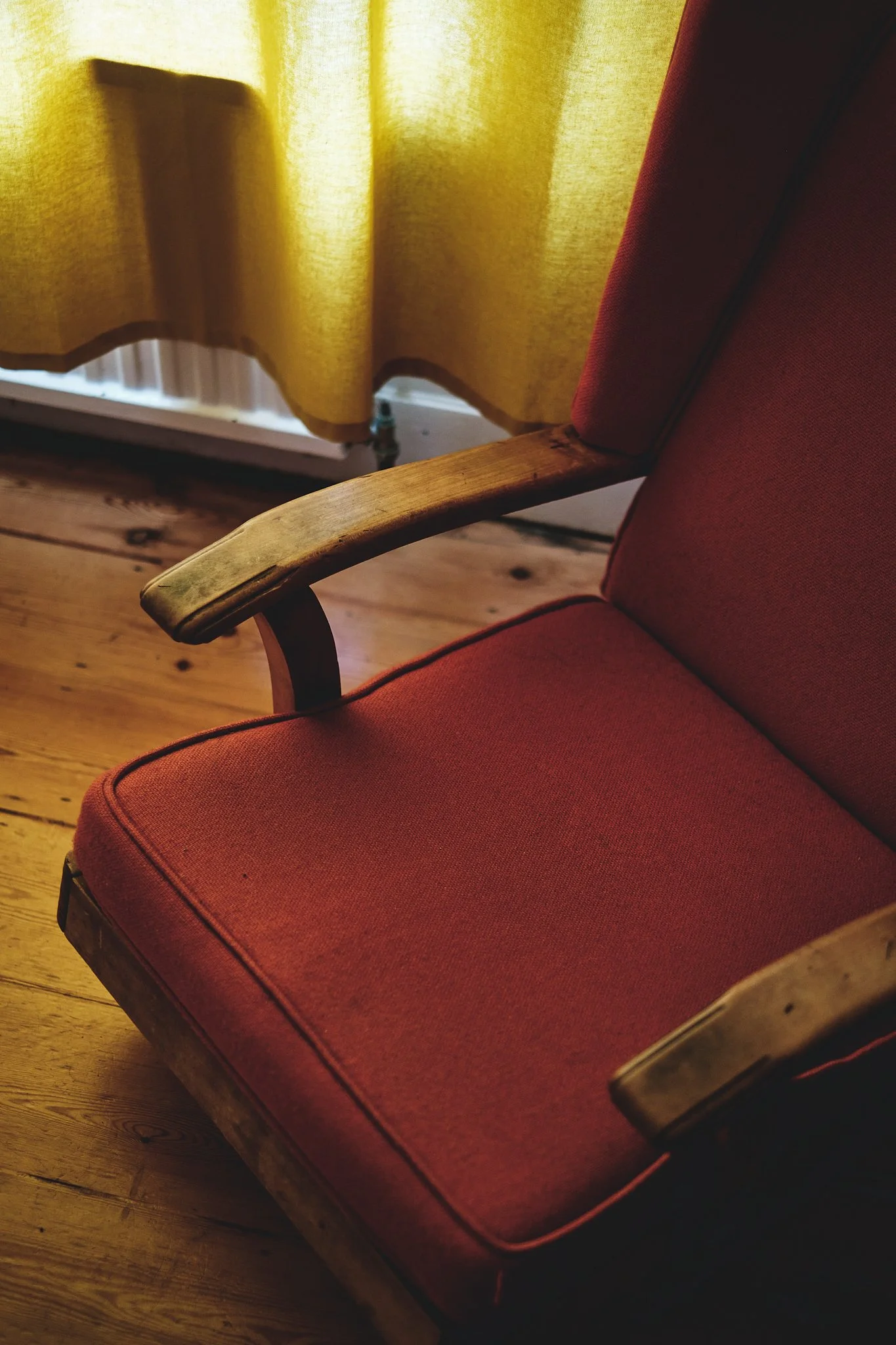 A vintage red upholstered armchair with wooden armrests in front of a yellow curtain and hardwood floor.