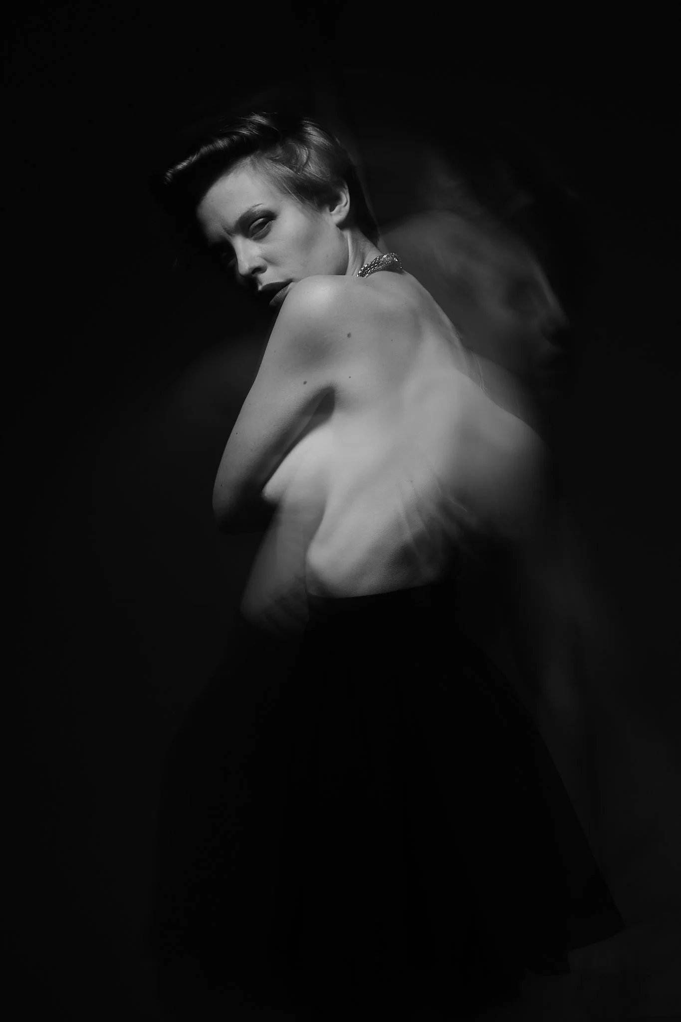 A black and white photograph of a woman with short dark hair, looking over her shoulder with a confident expression. The image has a blurred effect around her, creating a sense of motion.