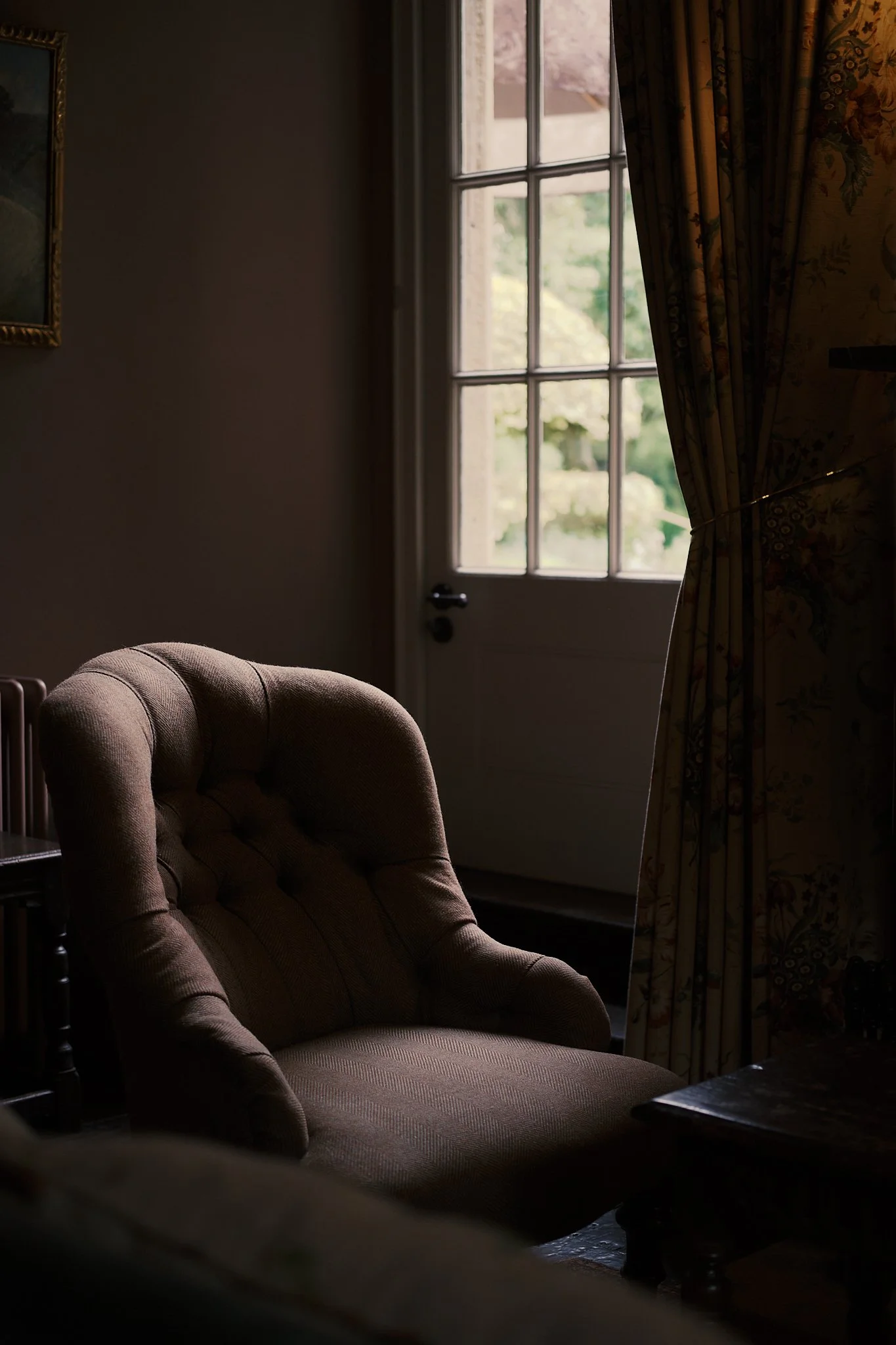 A brown upholstered armchair near a window with floral curtains, a door with glass panes, and a framed painting on the wall. Hotel interior photography at THE PIG-in the Cotswolds