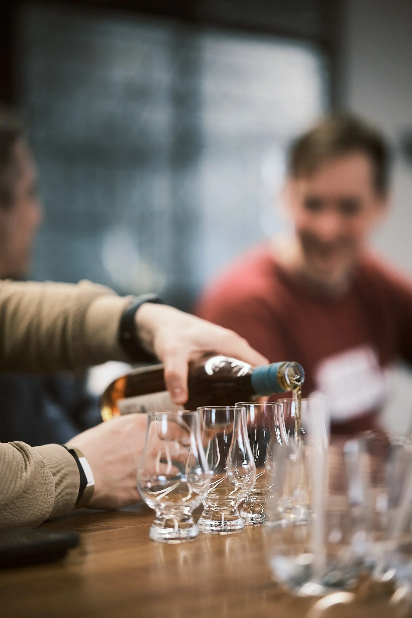 Person pouring a drink into small glasses on a wooden table, with another person smiling in the background.