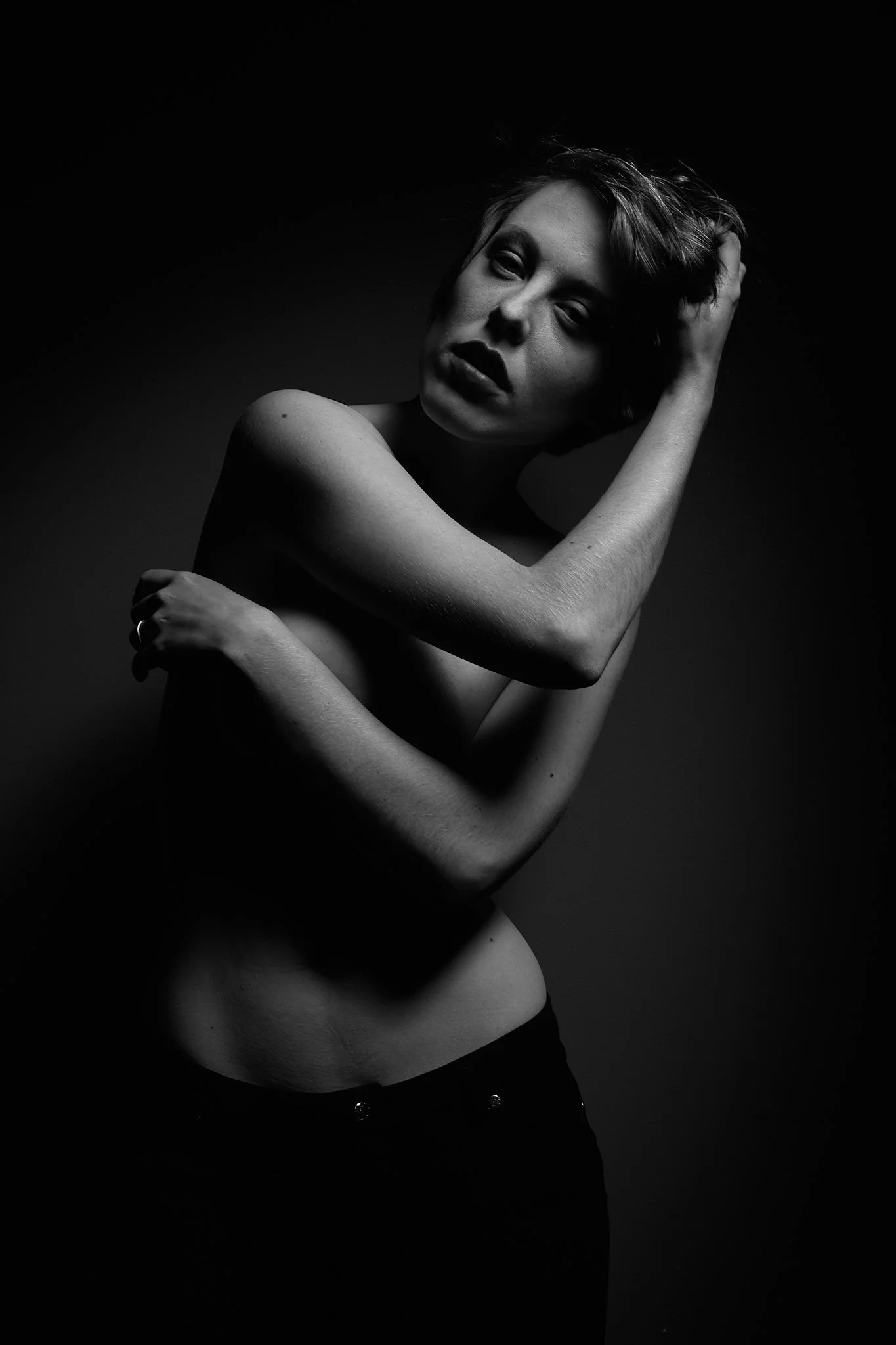 Black and white portrait of a topless woman with short hair, touching her head and posing against a dark background.