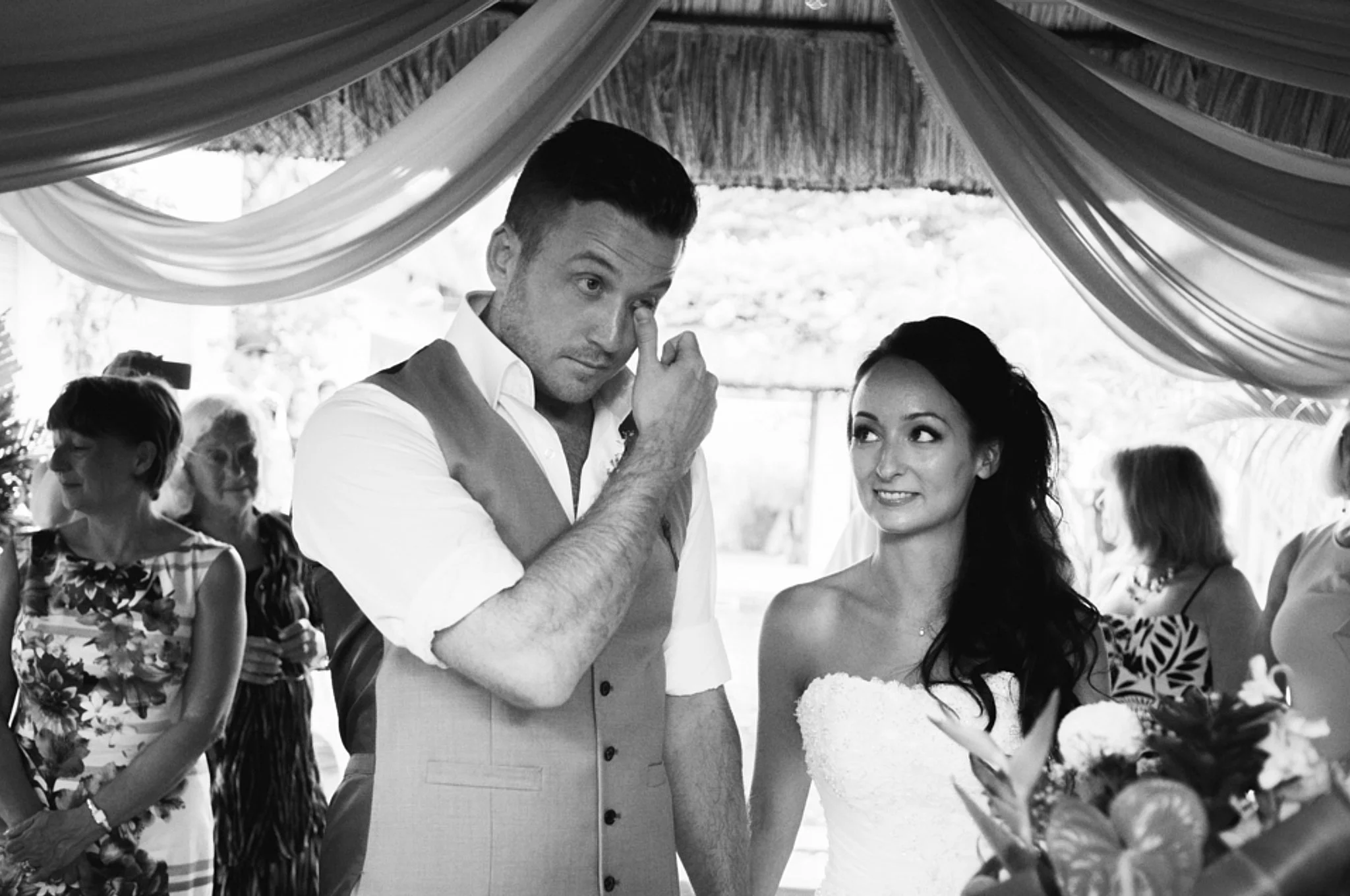 A man and woman at a wedding reception, standing under decorated canopy, with the man grooming his eye and the woman smiling, surrounded by other guests.