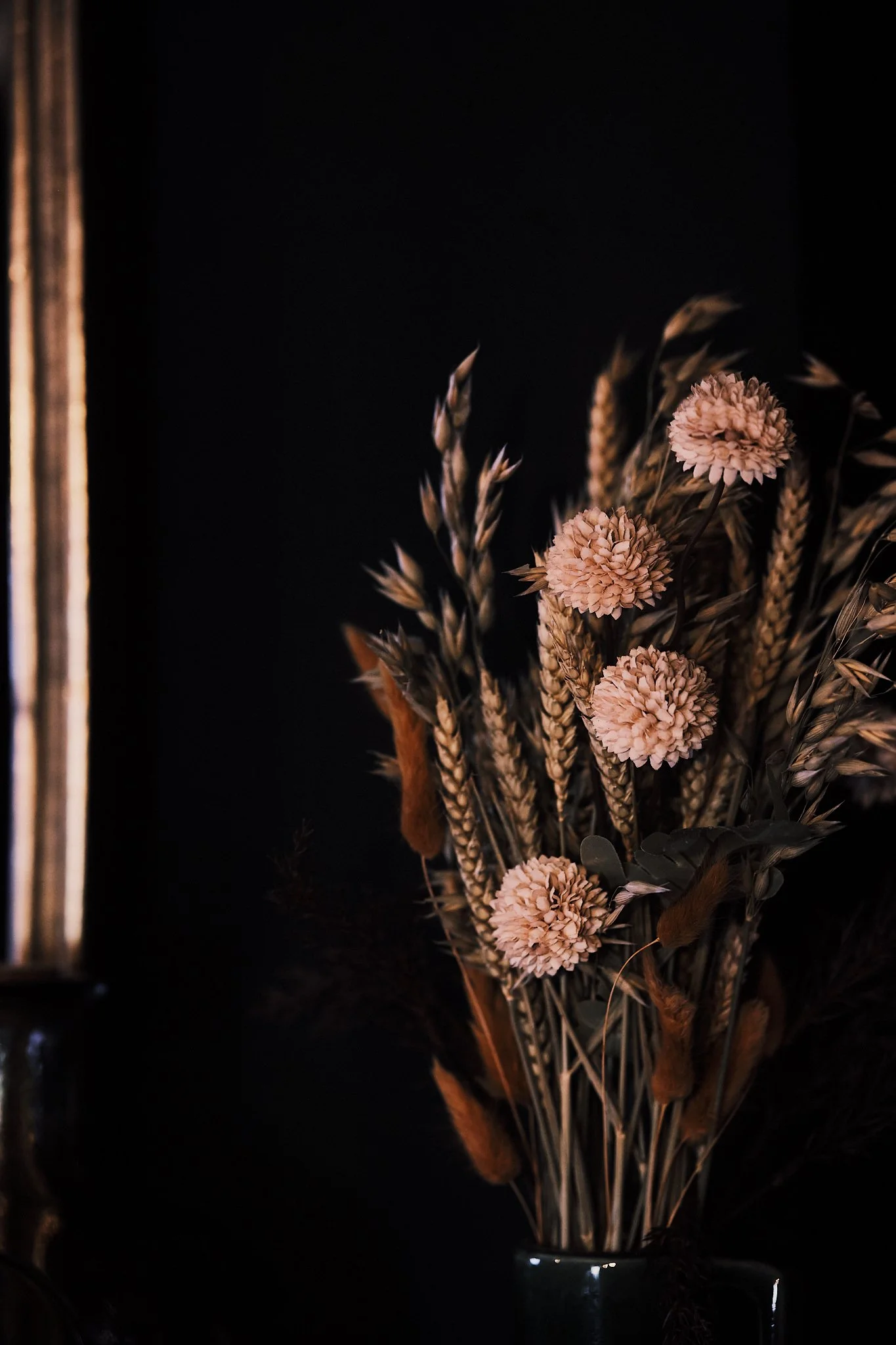 A dark-colored vase holding a bouquet of dried flowers, including pink pom-pom flowers, wheat, and other dried foliage, against a black background.