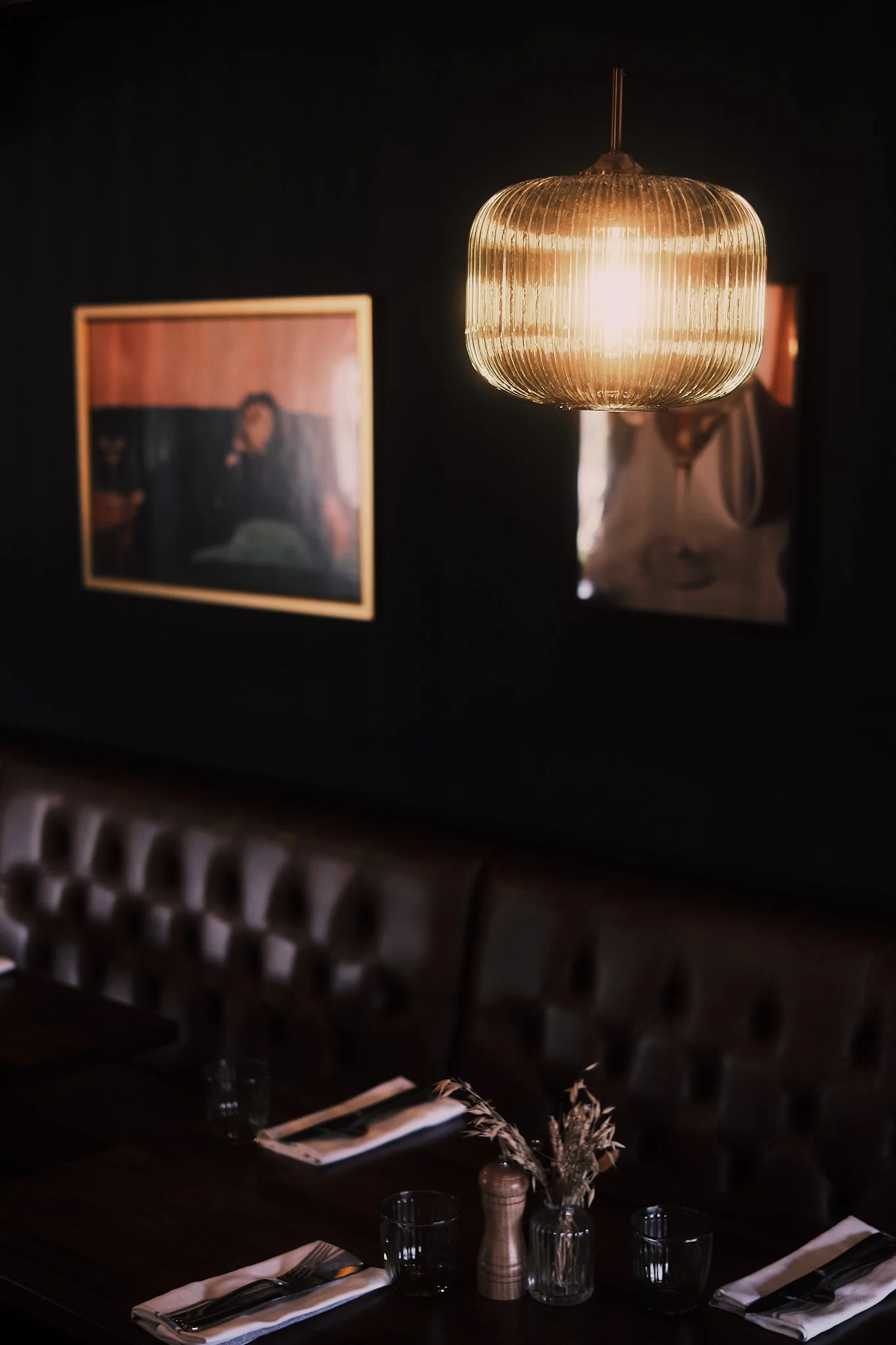 Interior of a restaurant with dim lighting, featuring a modern pendant light, framed artwork on a dark wall, a wooden table set with napkins, utensils, glasses, and a small flower arrangement.