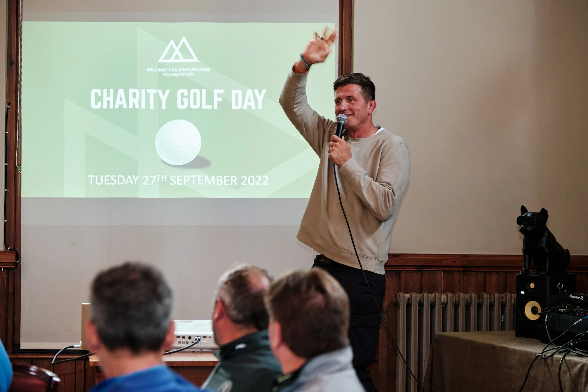A man speaking into a microphone with his hand raised, standing in front of a screen that reads 'Charity Golf Day, Tuesday 27th September 2022' at a charity event, with an audience seated in front of him.