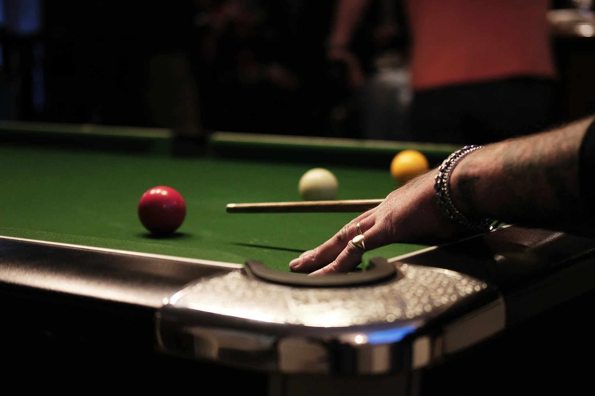 Close-up of a person's hand with rings and bracelets placing a cue stick on a snooker or pool table, with red, yellow, and white balls arranged on the green felt.