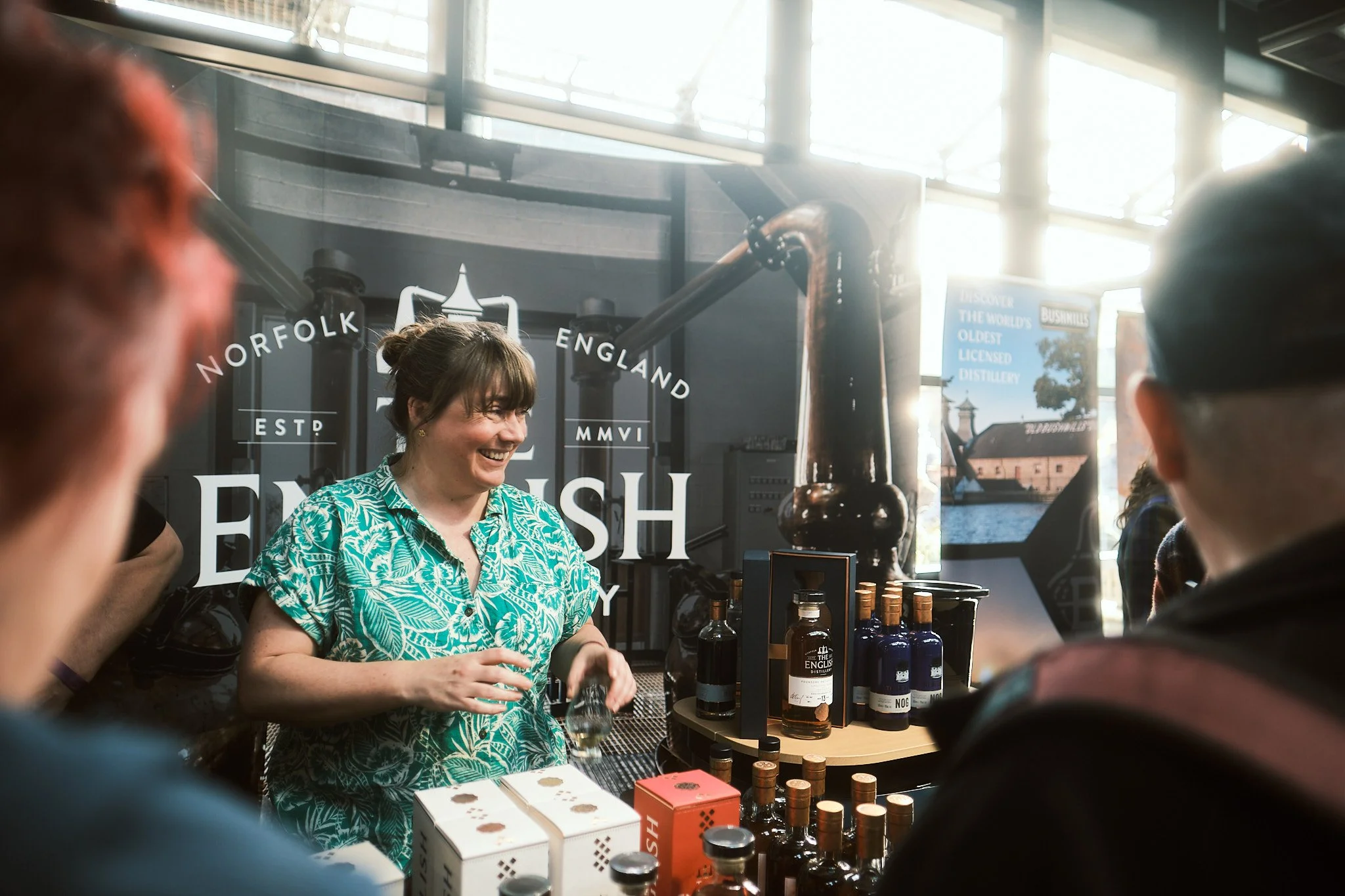 A woman in a blue patterned shirt is smiling and serving drinks at a distillery display. Several bottles of spirits are on the table, with a large distillation arm in the background. Two people are partially visible in the foreground, and a sign behi