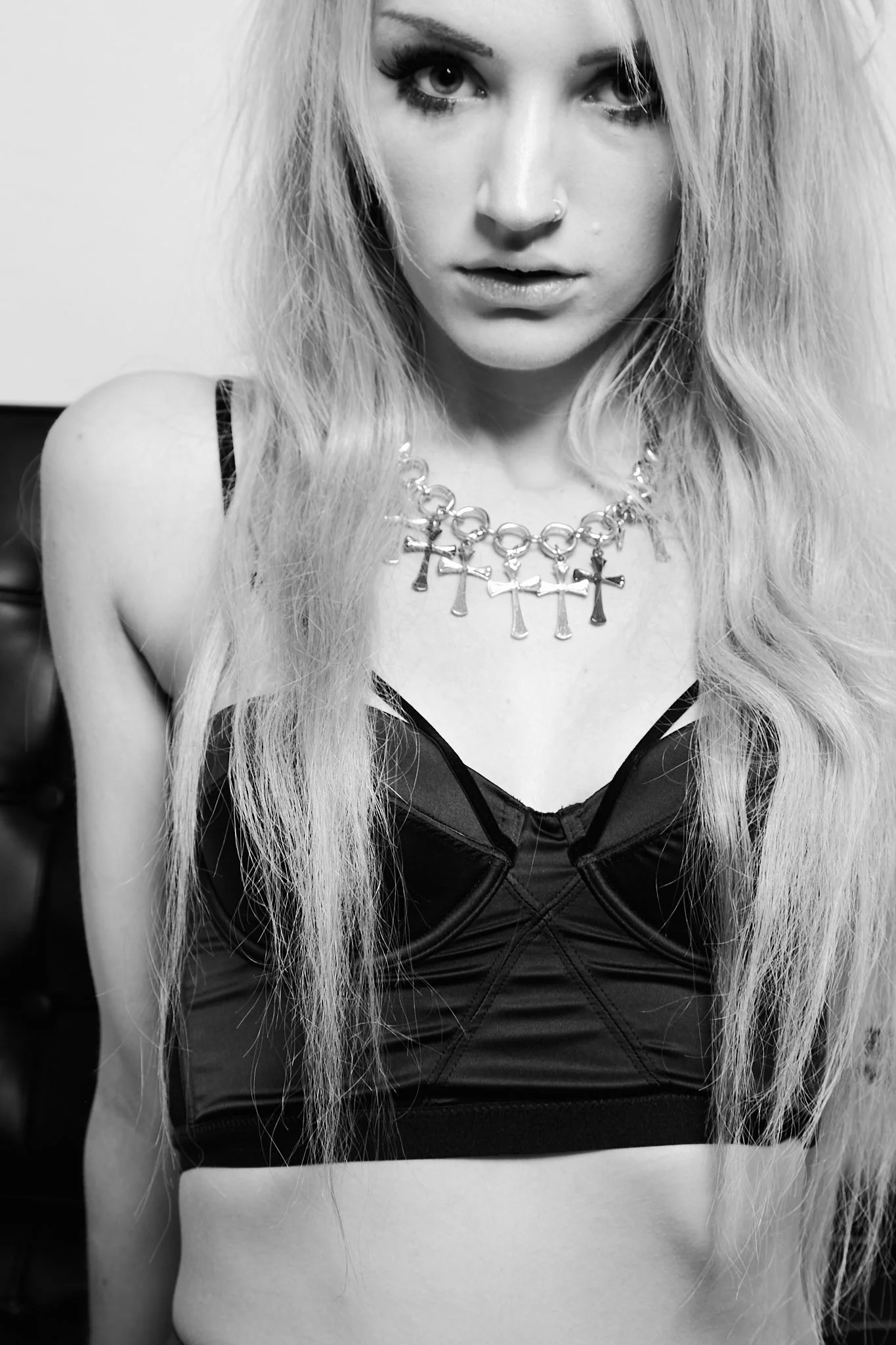 A black-and-white photo of a young woman with long, wavy blonde hair, wearing a necklace with multiple cross charms and a black top with thin straps, looking directly at the camera.