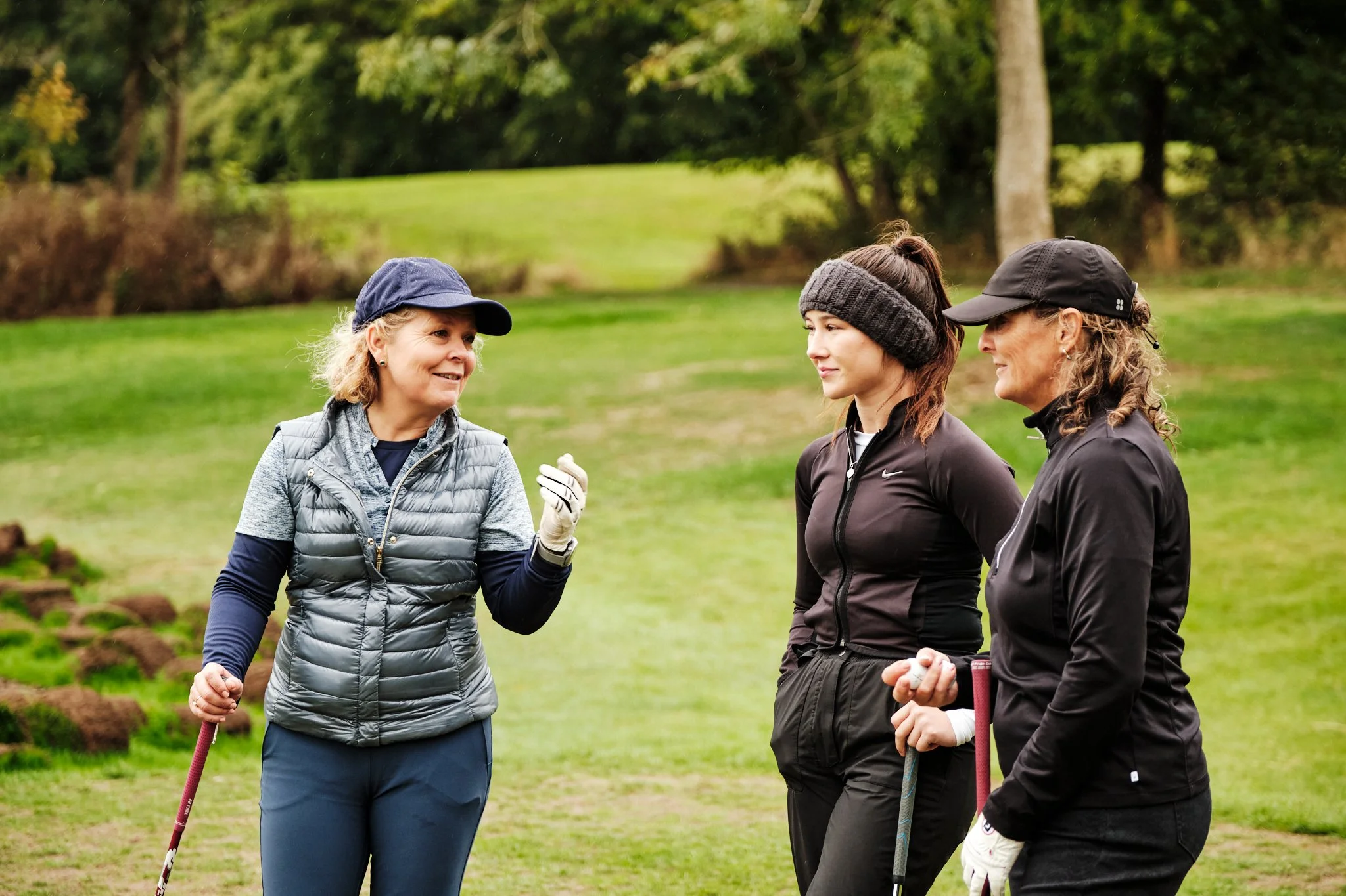 Three women dressed in golf attire standing on a golf course and having a conversation.