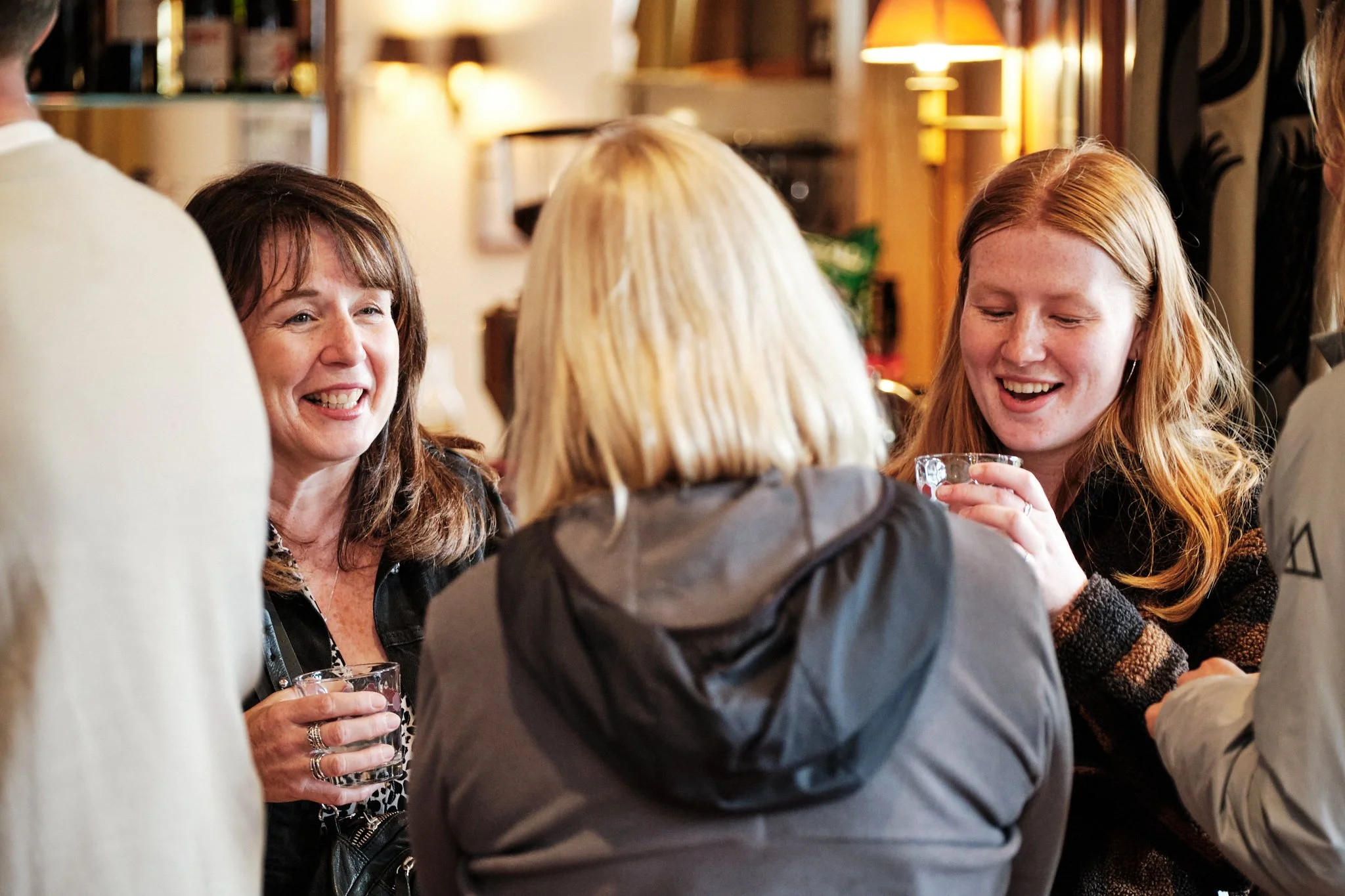 Four women gathered together, smiling and talking, holding drinks in a cozy, warmly lit indoor setting.