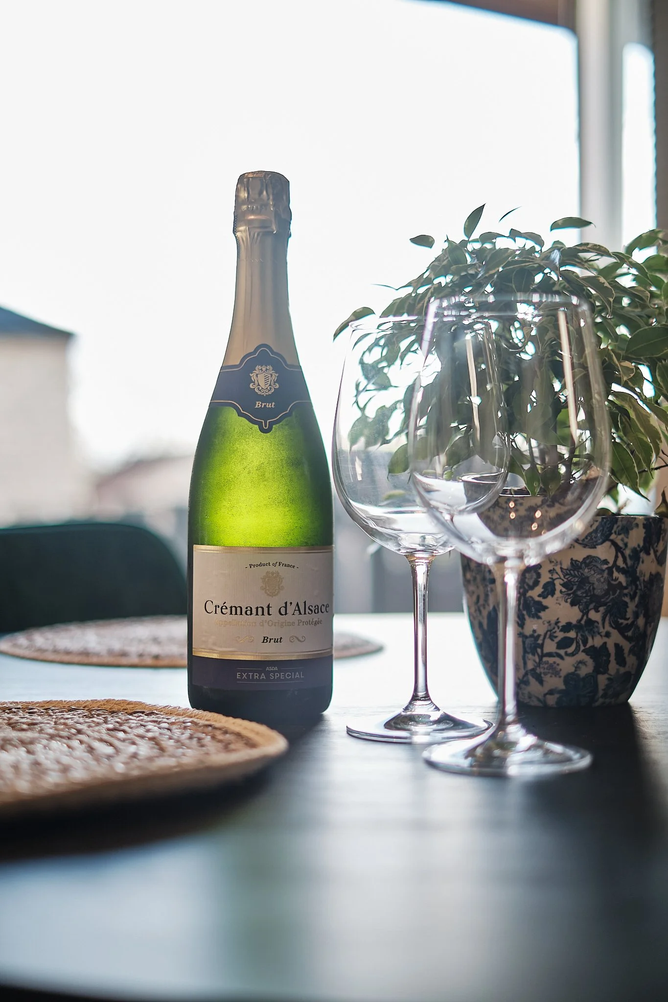 A bottle of Crémant d'Alsace sparkling wine, two empty wine glasses, and a potted plant on a dining table with woven placemats, set near a window with bright daylight.