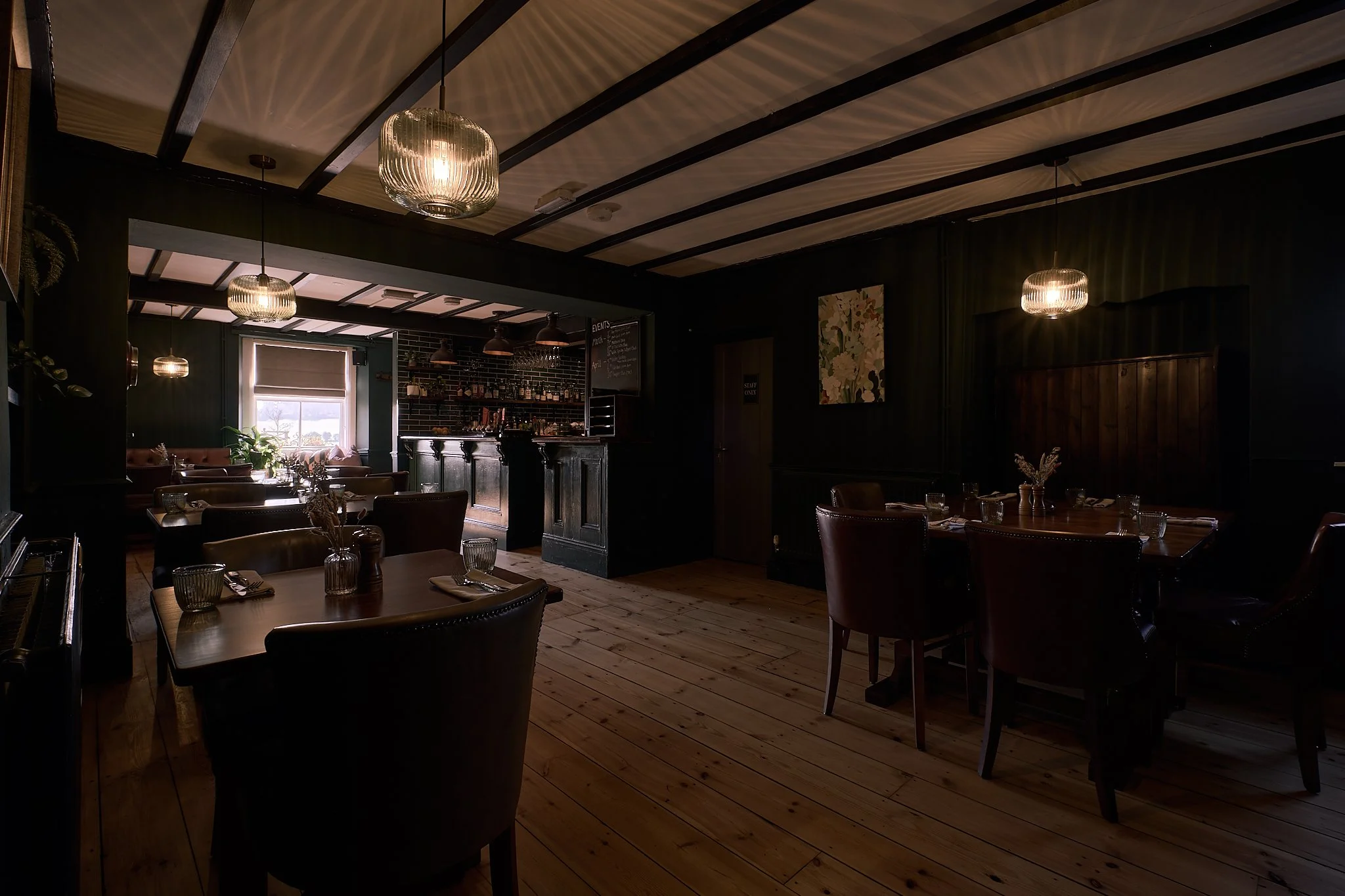 Dimly lit restaurant interior with wooden flooring, leather chairs, and hanging pendant lights, some tables set with glasses and utensils, a window letting in natural light, and a bar area in the background.