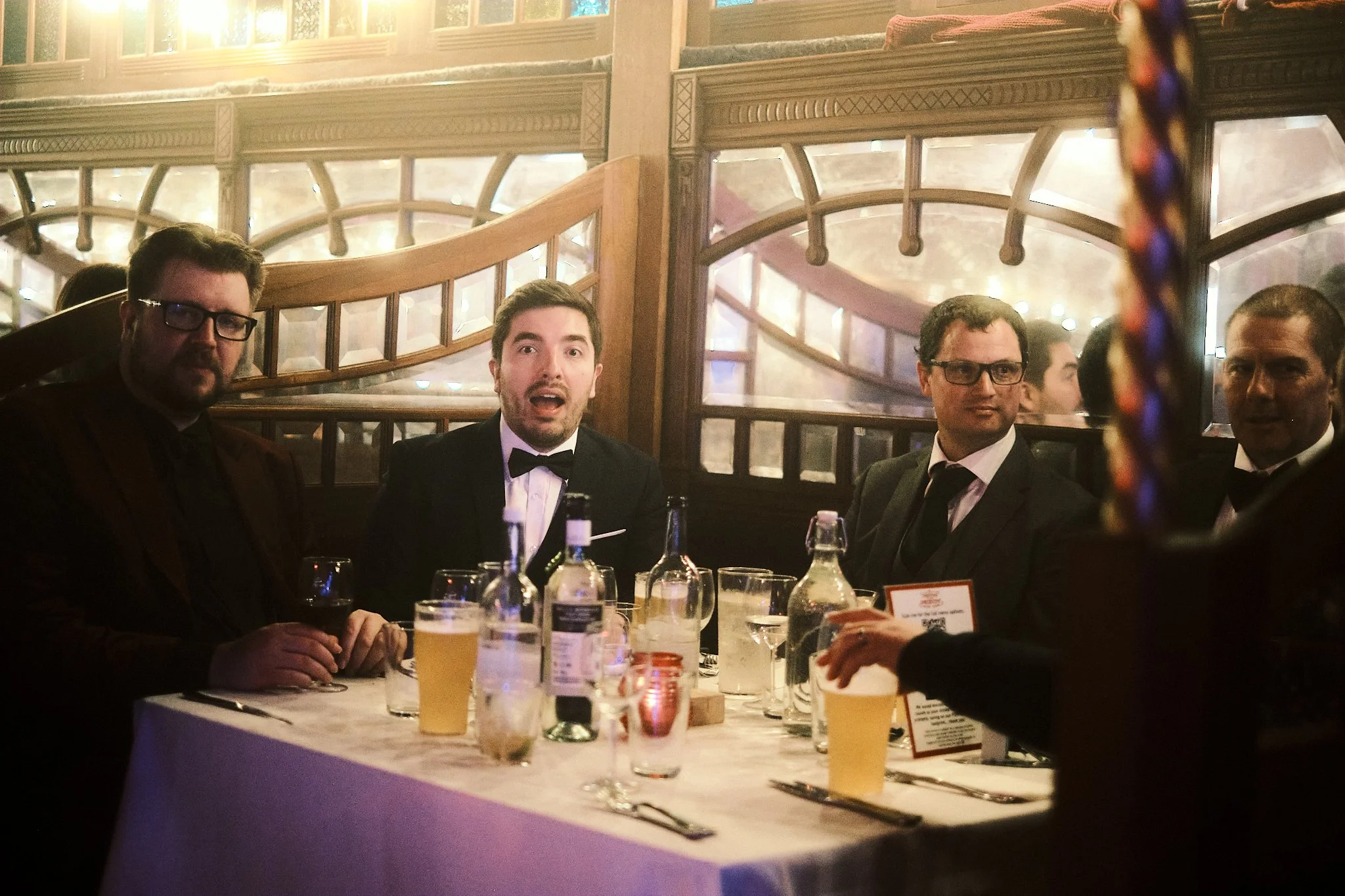 Four men in formal attire seated at a dinner table in an elegant restaurant, with drinks and bottles on the table, engaged in conversation.
