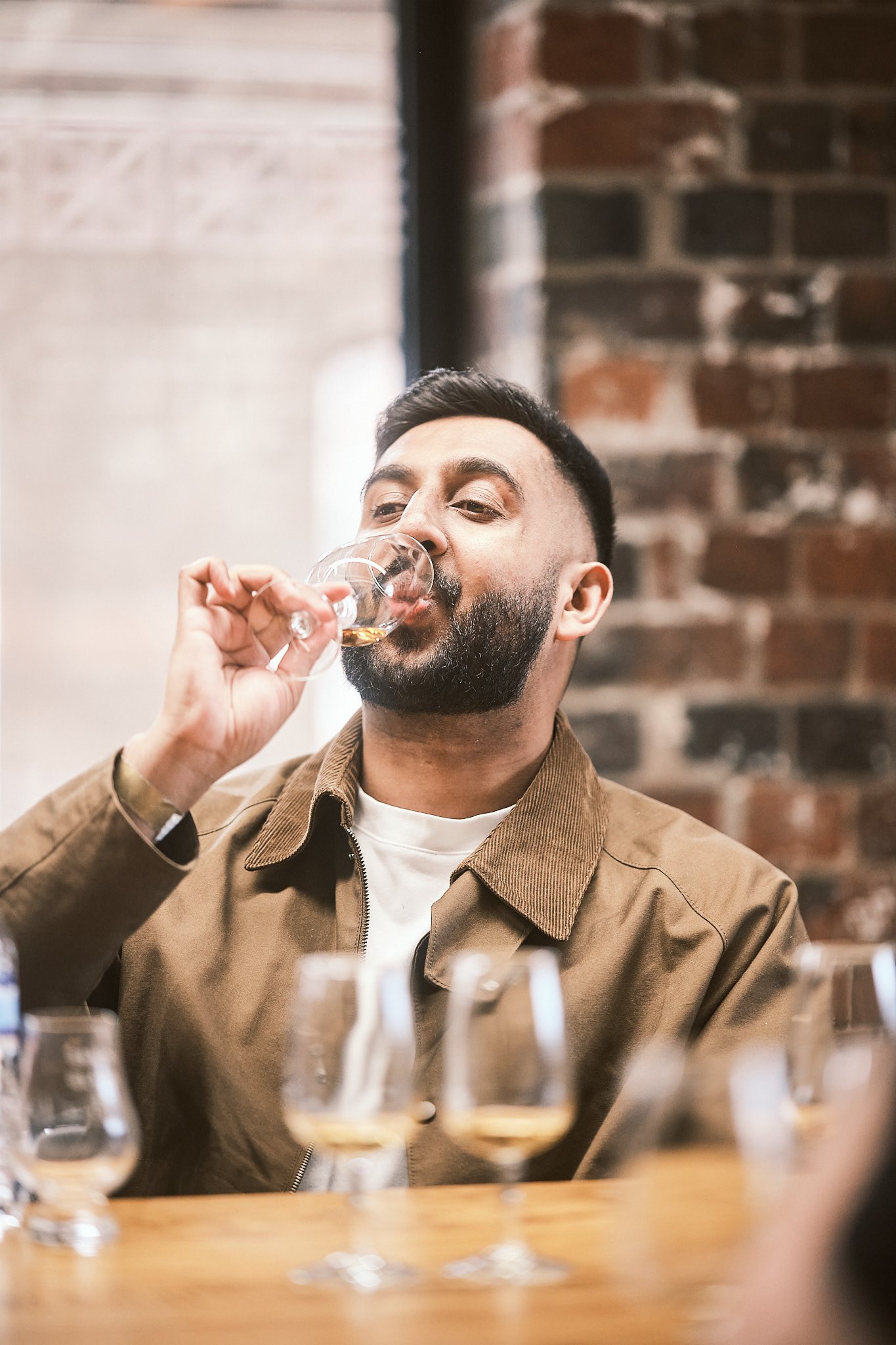 A man with a beard and short dark hair drinking from a glass of spirit, sitting at a table with several glasses of spirits in front of him, in a room with exposed brick walls.