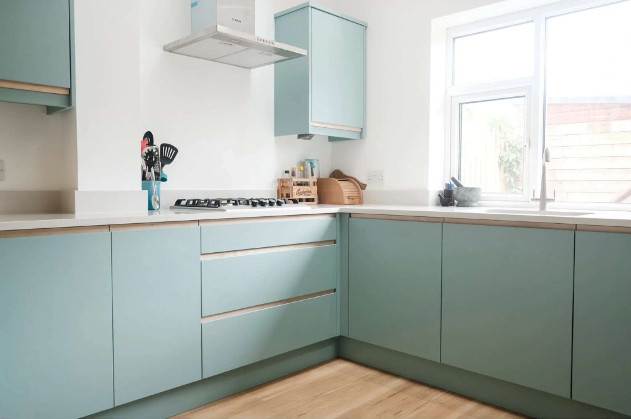 Light blue kitchen cabinets with white countertops, a gas stove, a window with a view of a wooden fence and some plants outside, and various kitchen utensils and items on the counters.