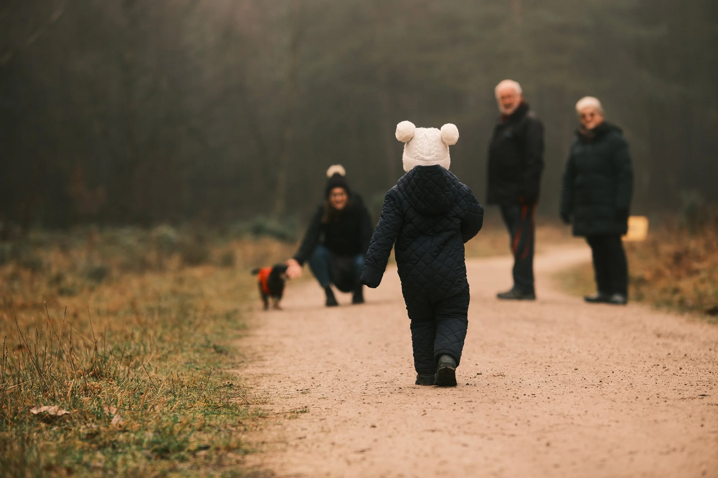 A young child wearing a white hat with pom-poms and a quilted black coat walking down a dirt path in a forest, with three adults and a dog in the background.