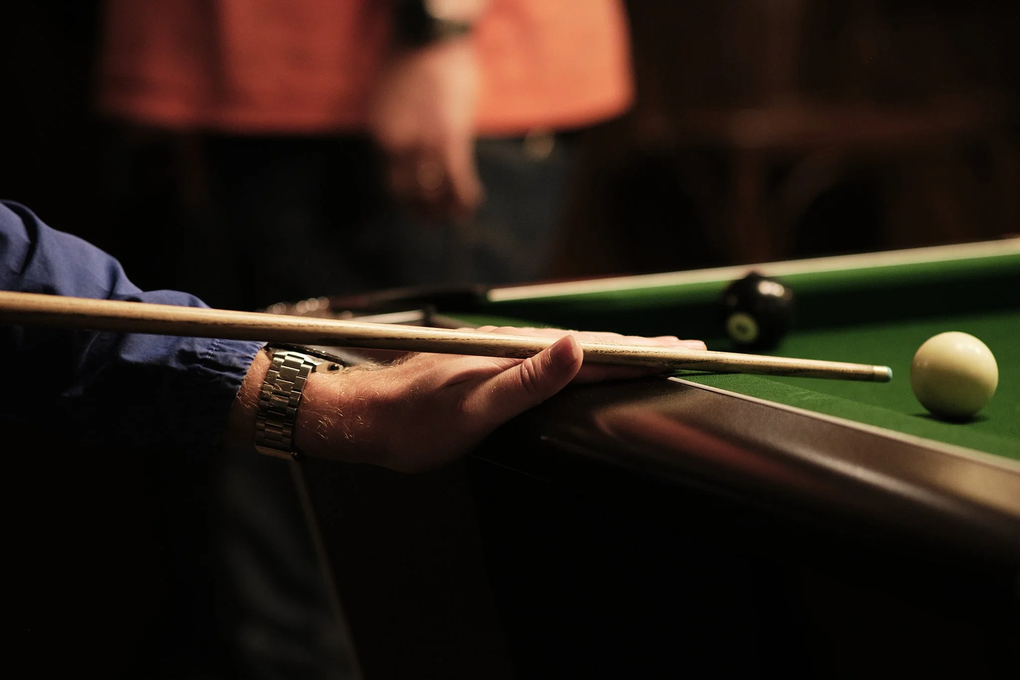 A person preparing to take a shot in a game of billiards or pool, holding a cue stick with their left hand on the edge of the table, with a white cue ball in position on the green felt pool table.