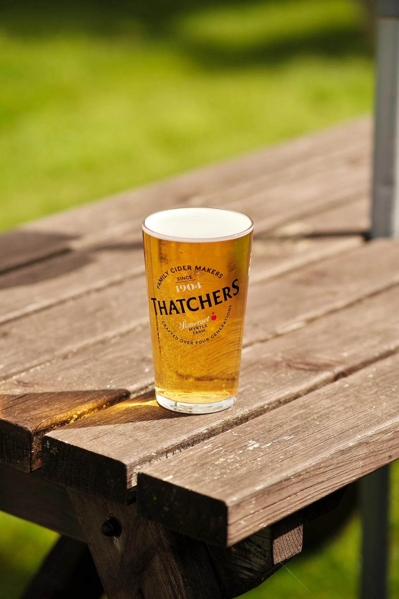 A pint glass of golden beer with a white head on a wooden picnic table outdoors.