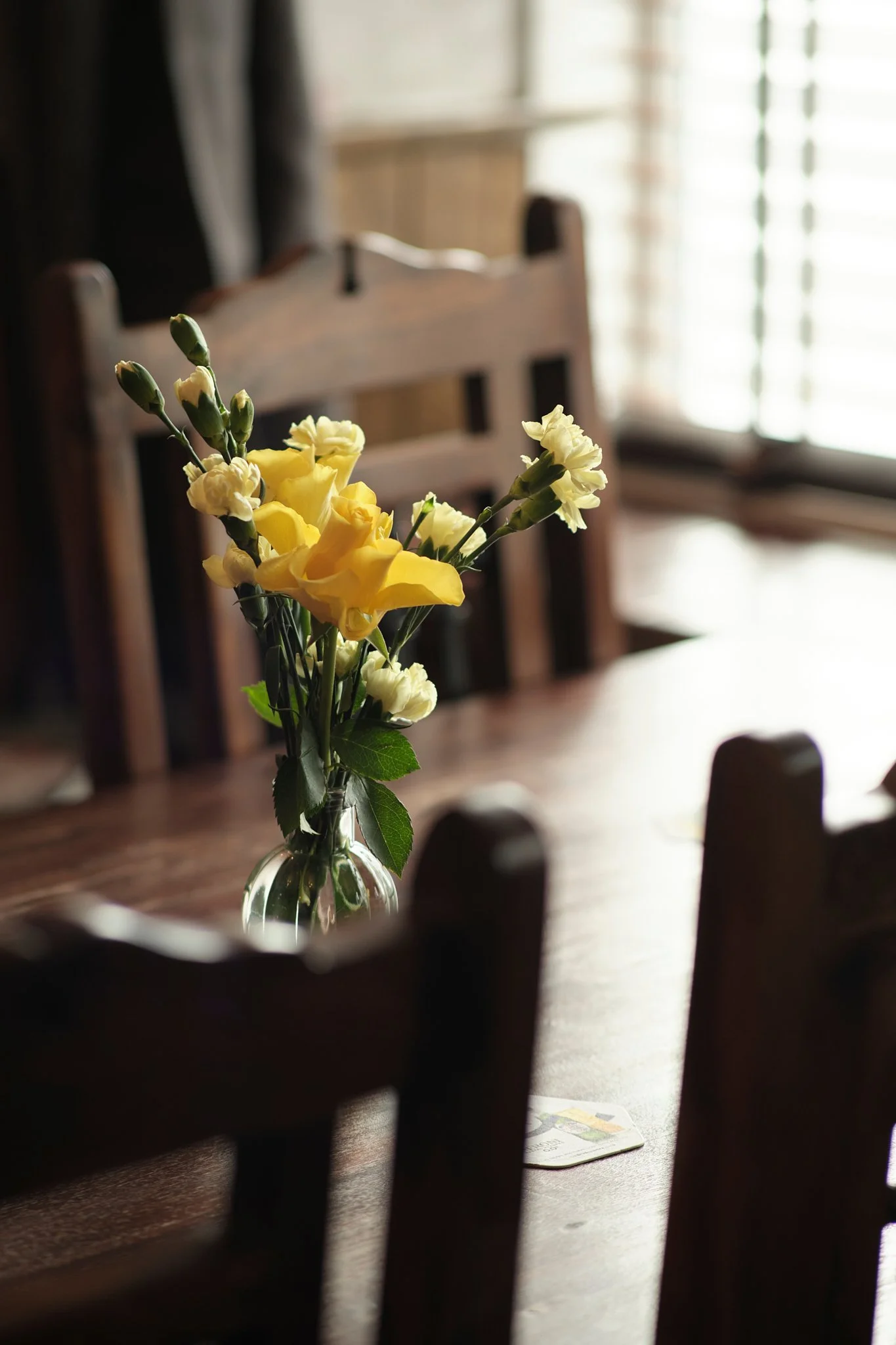 A glass vase with yellow and white flowers on a wooden dining table in a warmly lit room.