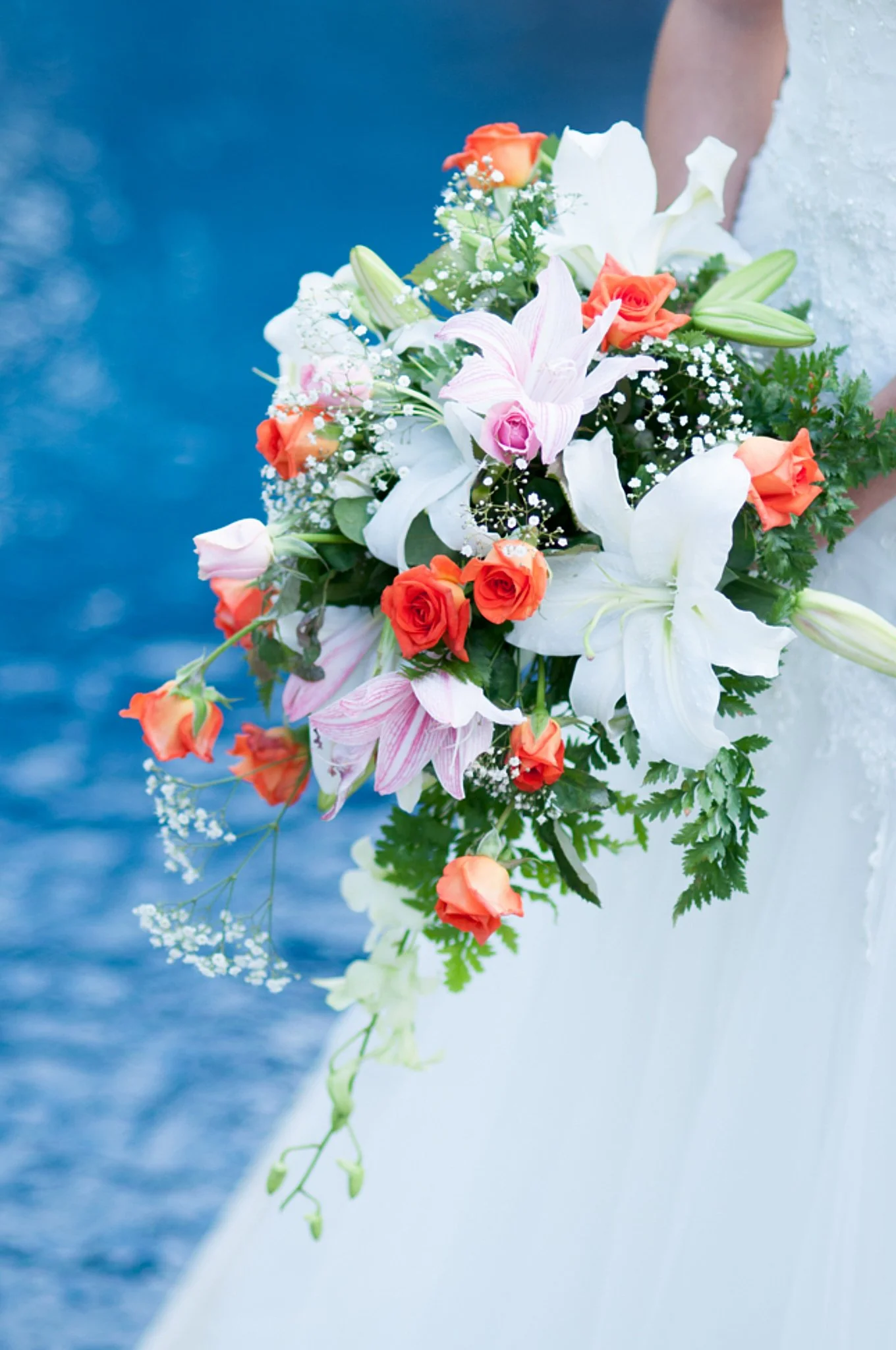 Close-up of a bride holding a cascading bouquet of white lilies, pink roses, orange roses, and baby's breath against a blurred blue background.
