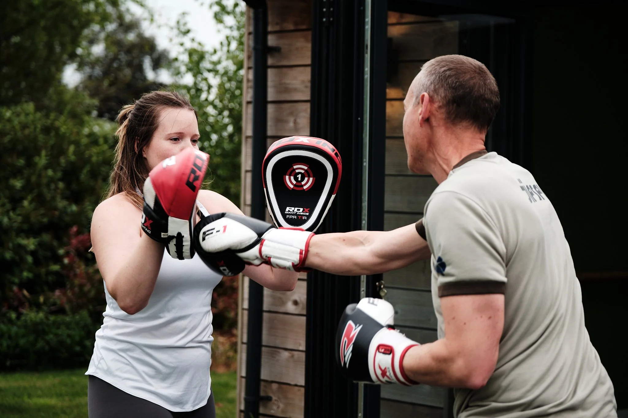 A young woman practicing boxing with a coach outdoors, wearing boxing gloves and focusing on a punching pad held by the coach near a wooden building with greenery in the background.