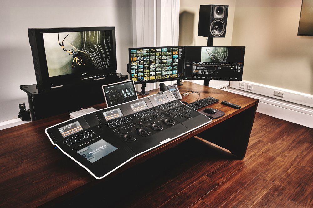 A professional audio and video editing workspace with three monitors, a mixing console, computer peripherals, a speaker, and a television in a room with wood flooring.