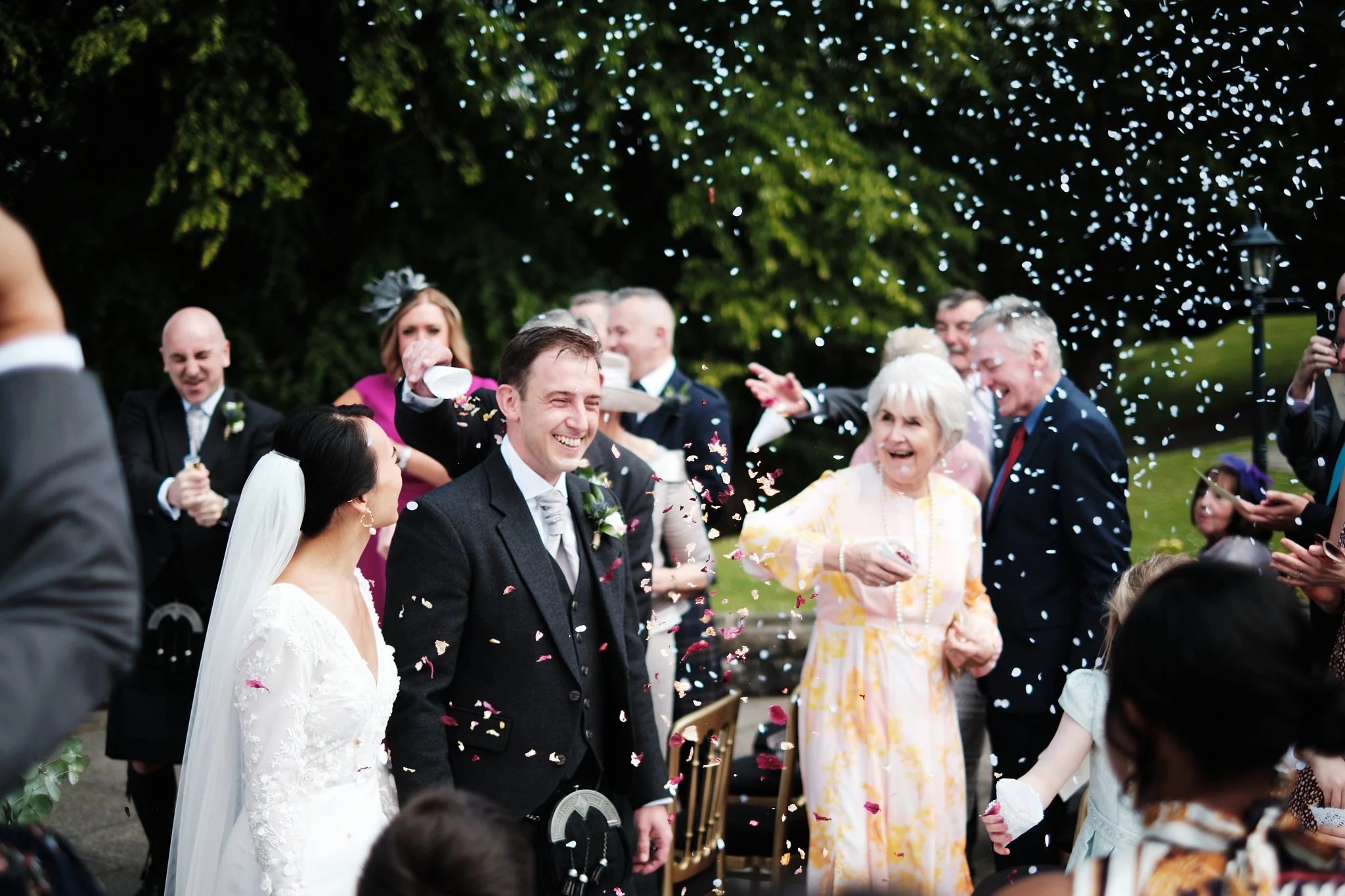 Wedding celebration outdoors with guests throwing colorful confetti, smiling, and enjoying the event under a canopy of string lights.