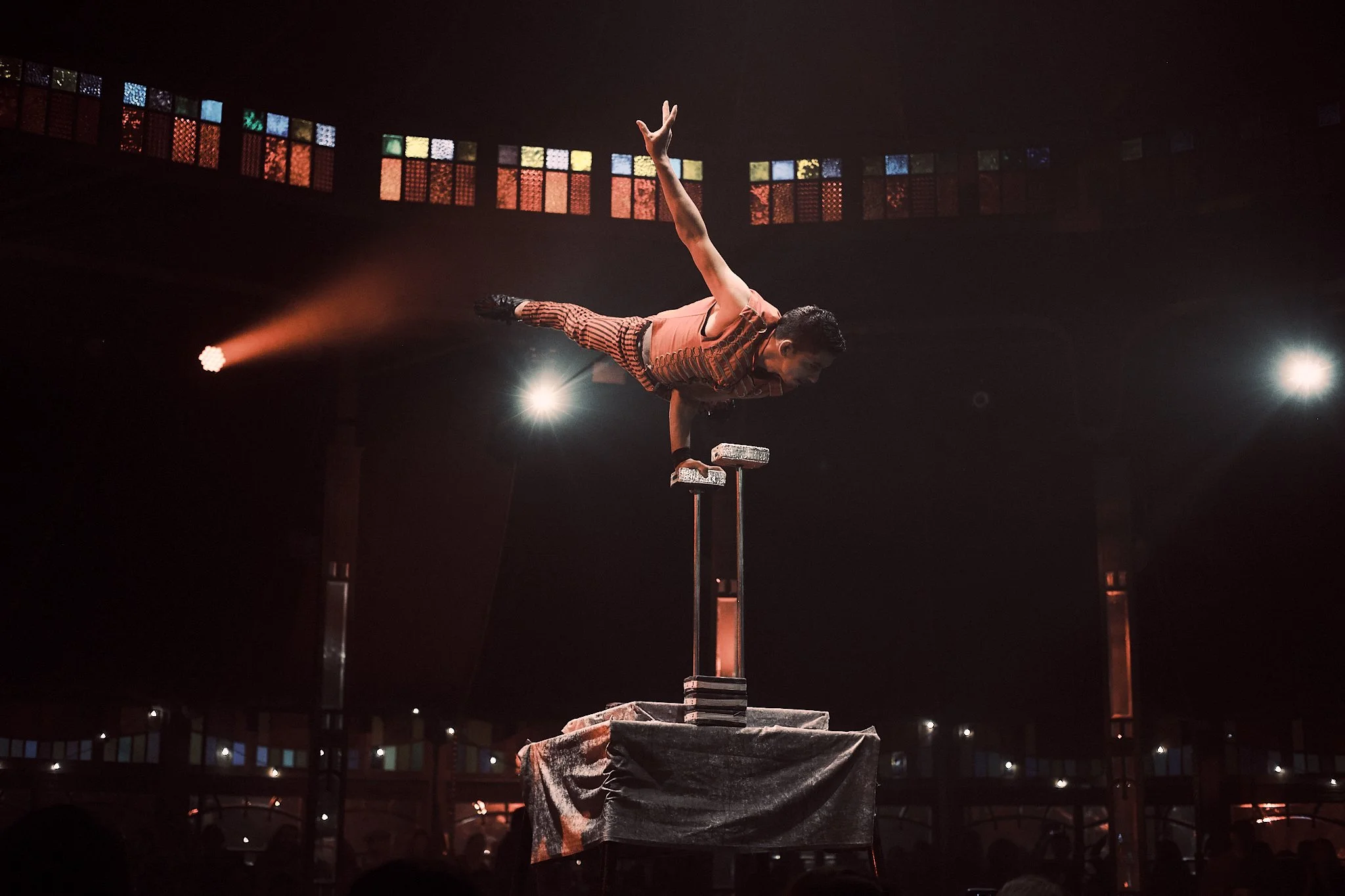 A performer in striped clothing balancing on one hand on a stack of blocks during a balancing act in a dark theater with colorful lights and spotlights.