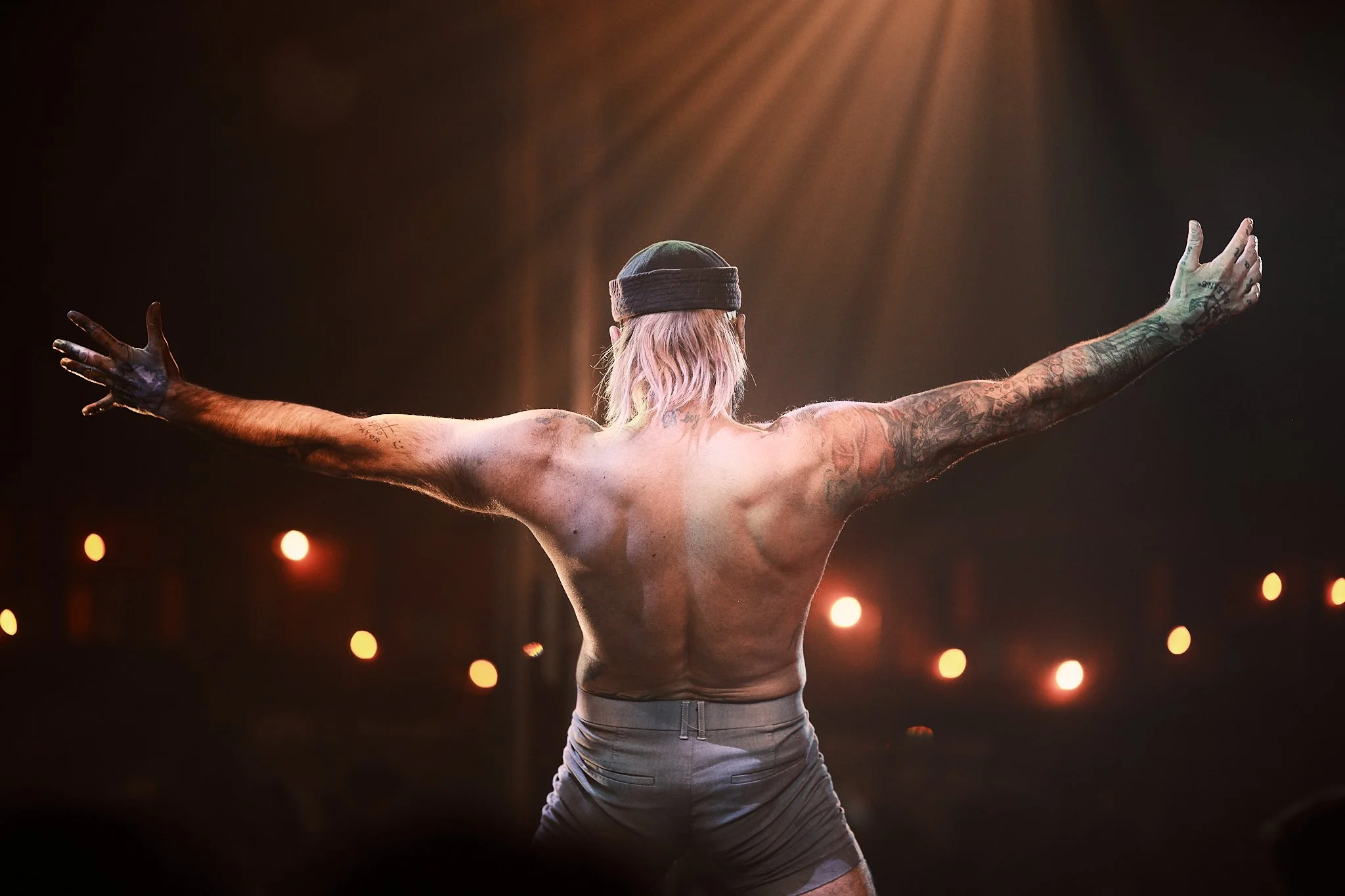 A shirtless performer with tattoos, wearing a headband and shorts, on stage with arms wide open, back facing the camera, stage lights shining down.