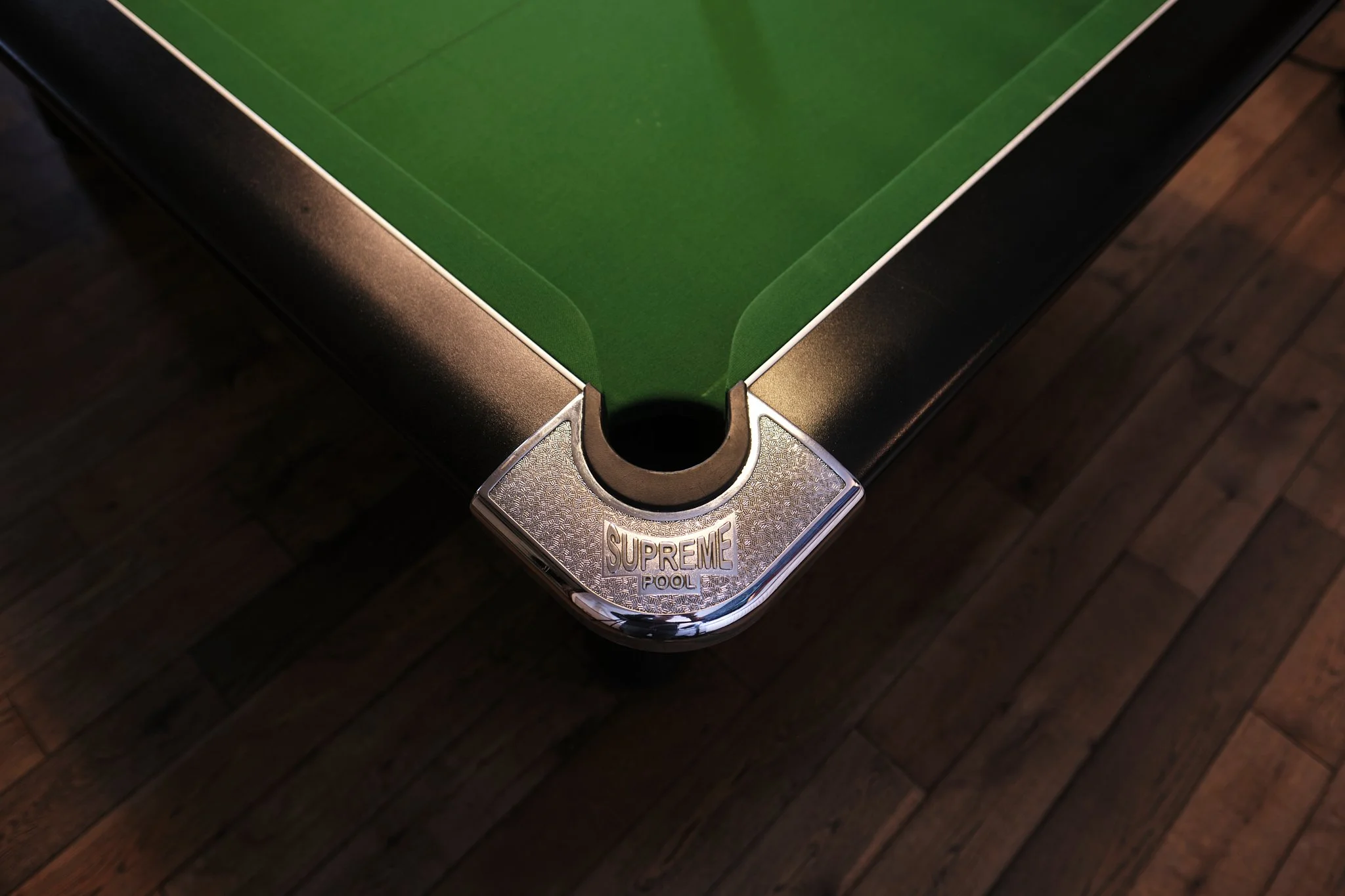 Close-up of a corner of a pool table with a green playing surface and a metal corner with the label 'Supreme Pool', on a wooden floor.