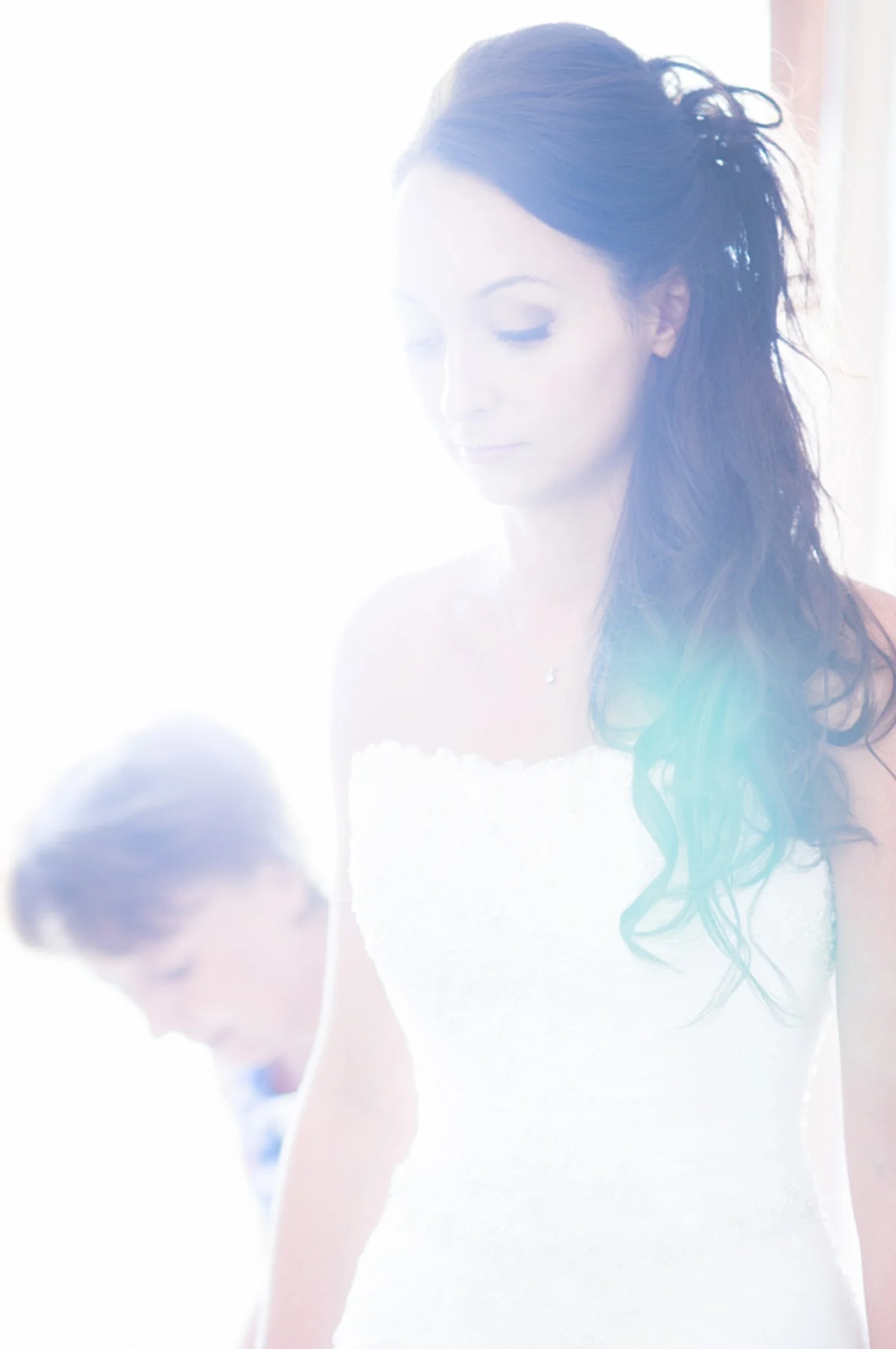 A woman with dark, curly hair and light makeup, wearing a white dress, standing near a window with bright sunlight, looking down.