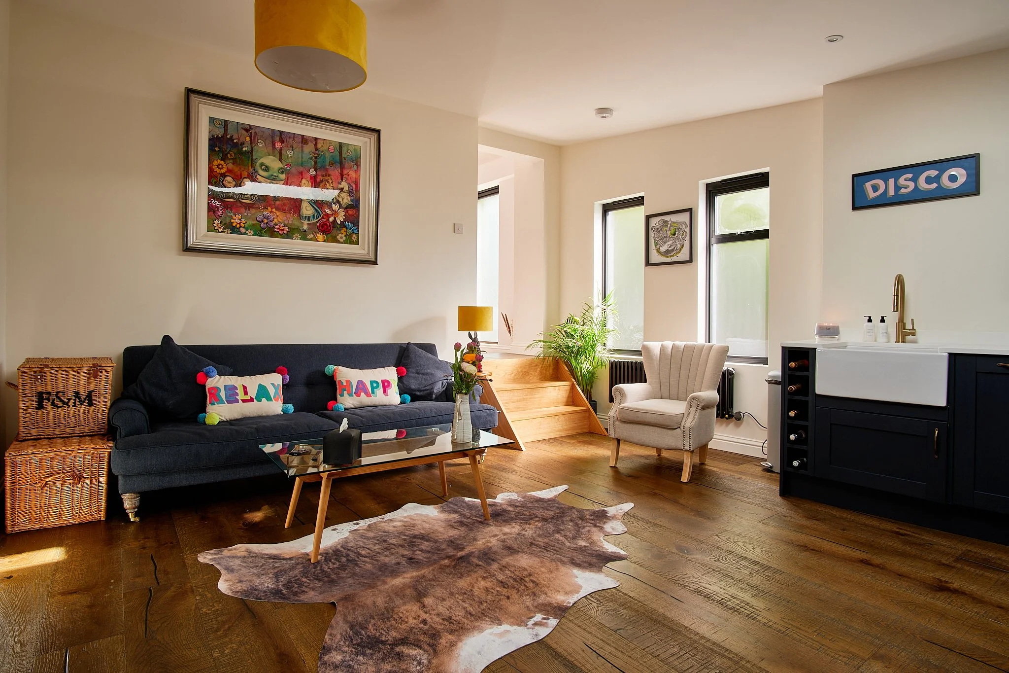 Living room with a dark blue sofa, colorful pillows, a glass coffee table, cowhide rug, white armchair, indoor plants, wooden stairs, large windows, and decorative wall art.