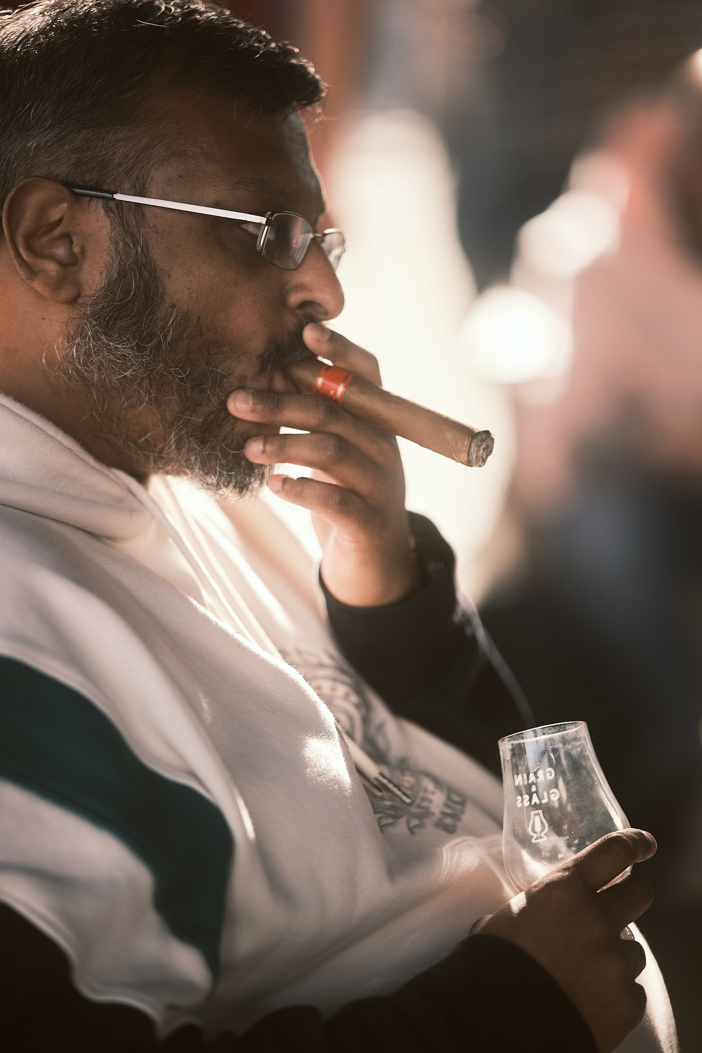 Man with glasses and a beard smoking a cigar, holding a glass, in a casual setting.