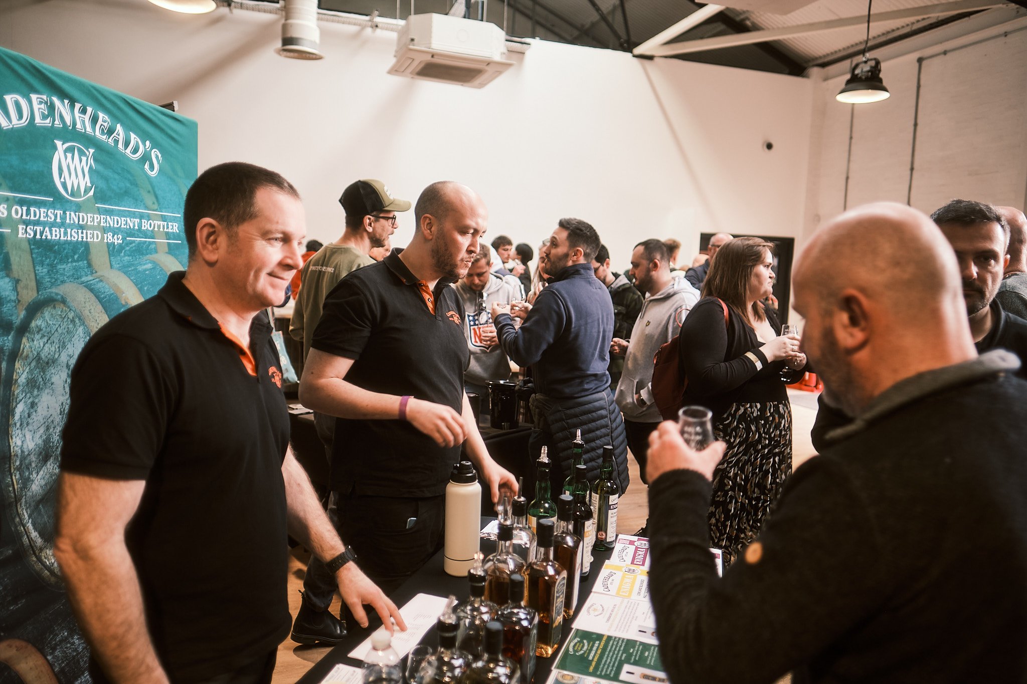 People attending a whiskey tasting event, sampling drinks and engaging in conversations, with bottles of whiskey on the table and a green banner in the background.