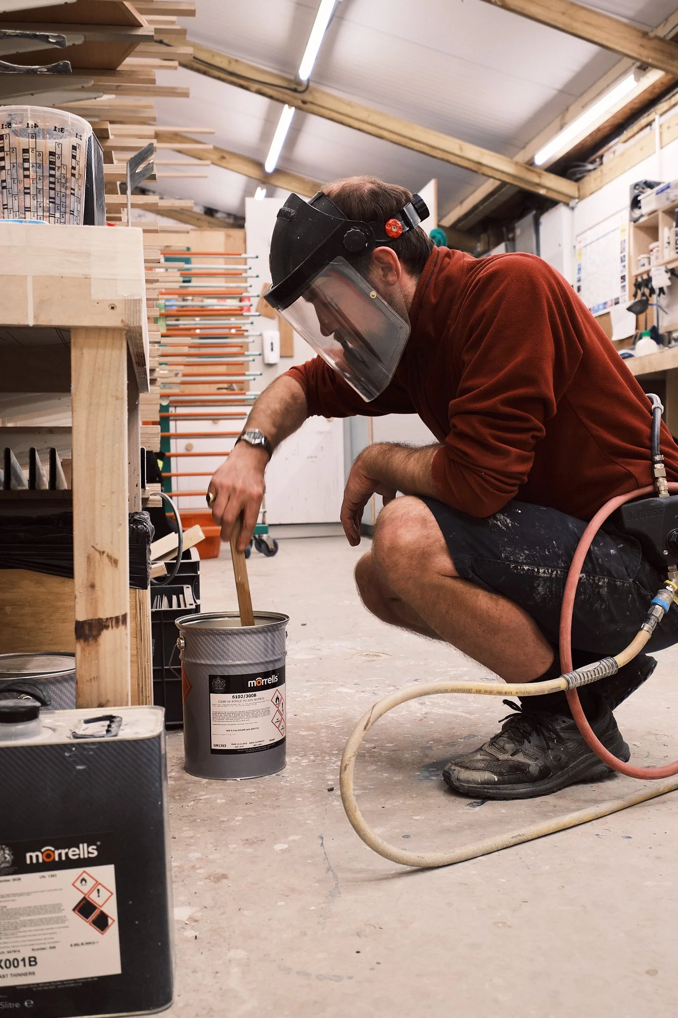 A man wearing a welding helmet and safety gear kneels on the floor of a woodworking workshop, stirring a can of paint or lacquer with a wooden stick.