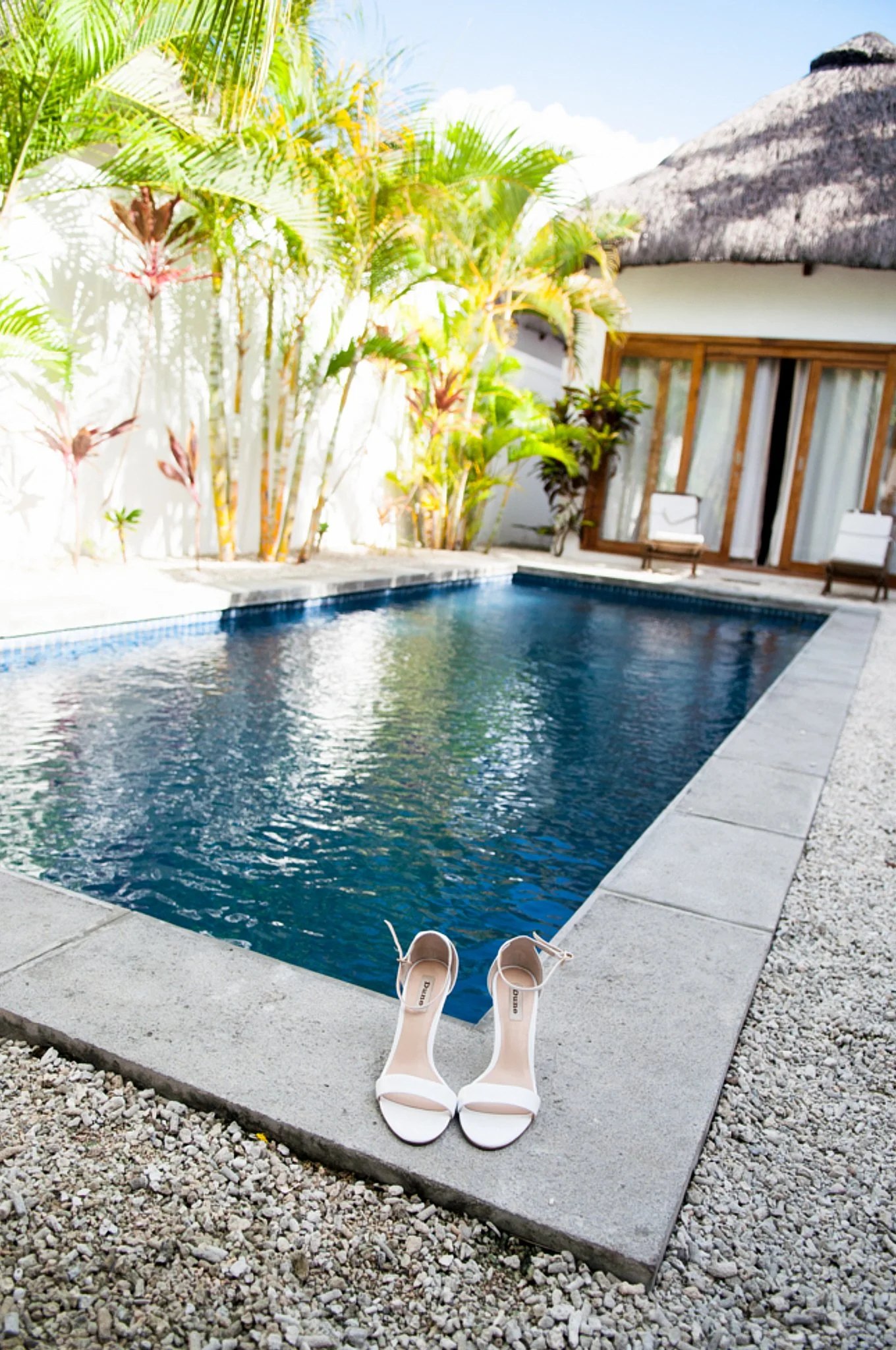 A pair of white high-heeled shoes placed on the edge of a swimming pool patio, with a view of a blue pool, tropical plants, a thatched roof building, and a seating area with chairs and curtains in the background.