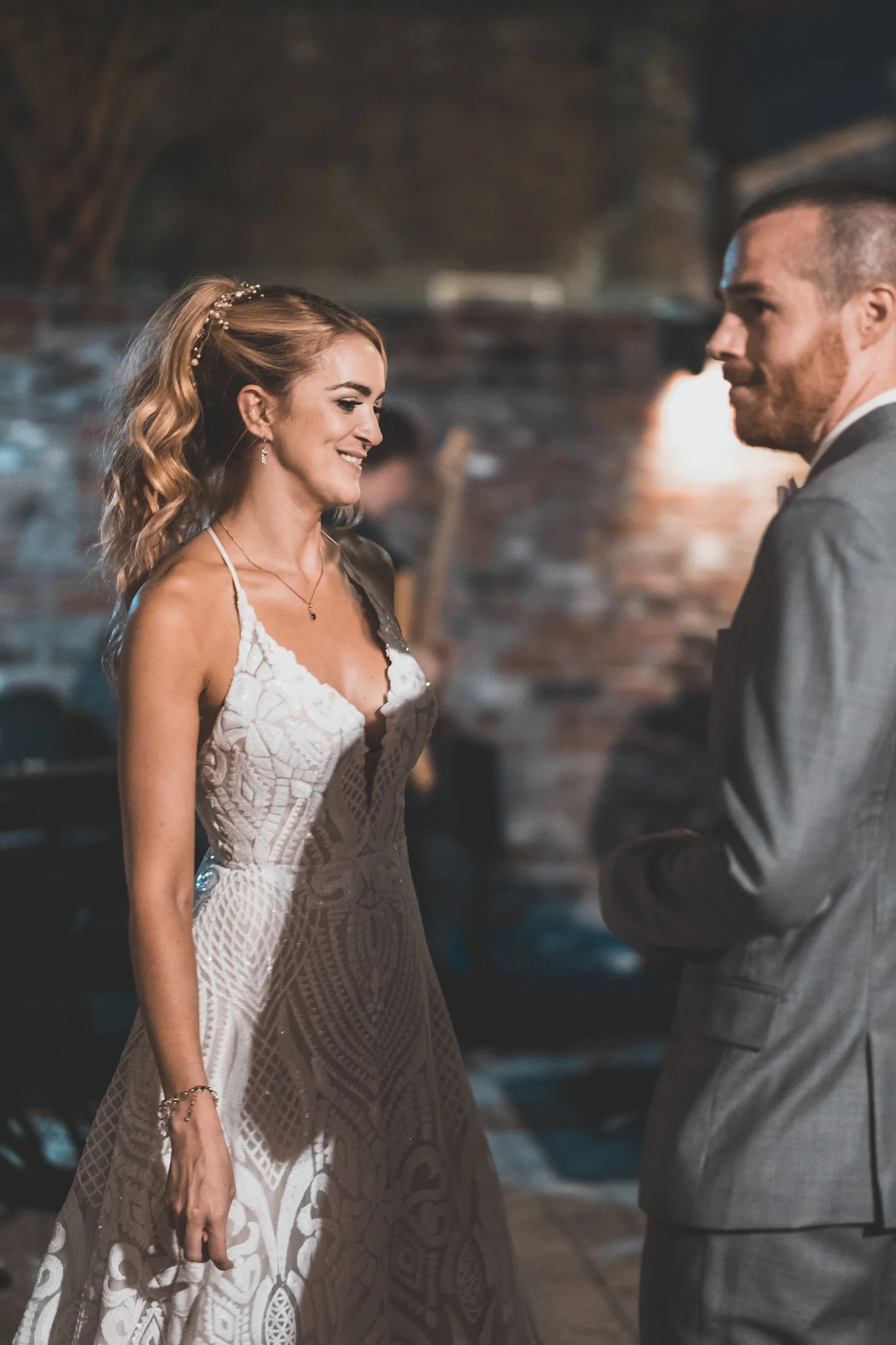 A woman in a white sleeveless lace dress smiling at a man in a gray suit with a bow tie during a wedding or formal event, with a blurred background of brick walls and warm lighting.