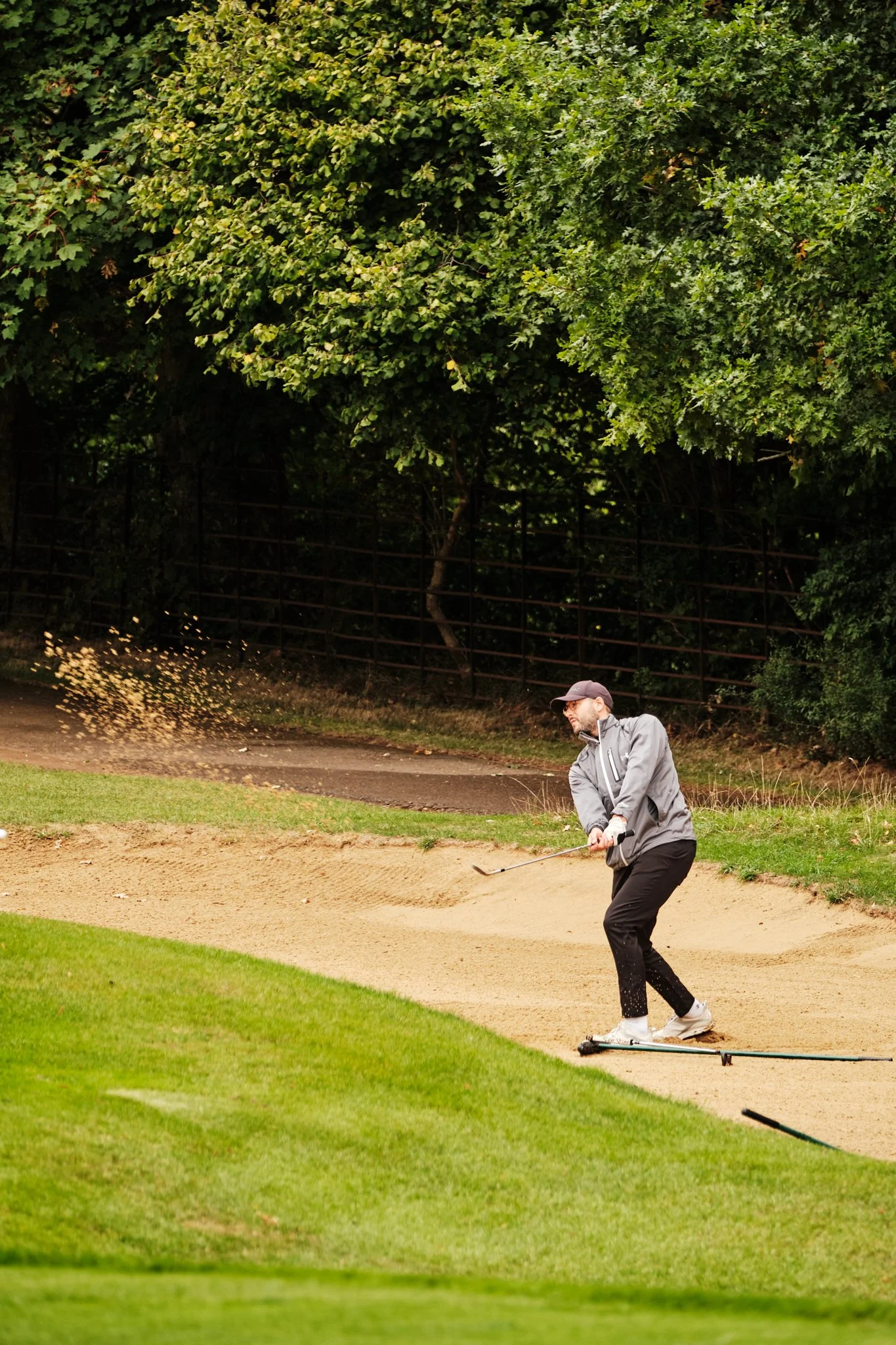 A man in a gray jacket and gray cap is hitting a golf ball out of a sand bunker on a golf course, with trees and a fence in the background.