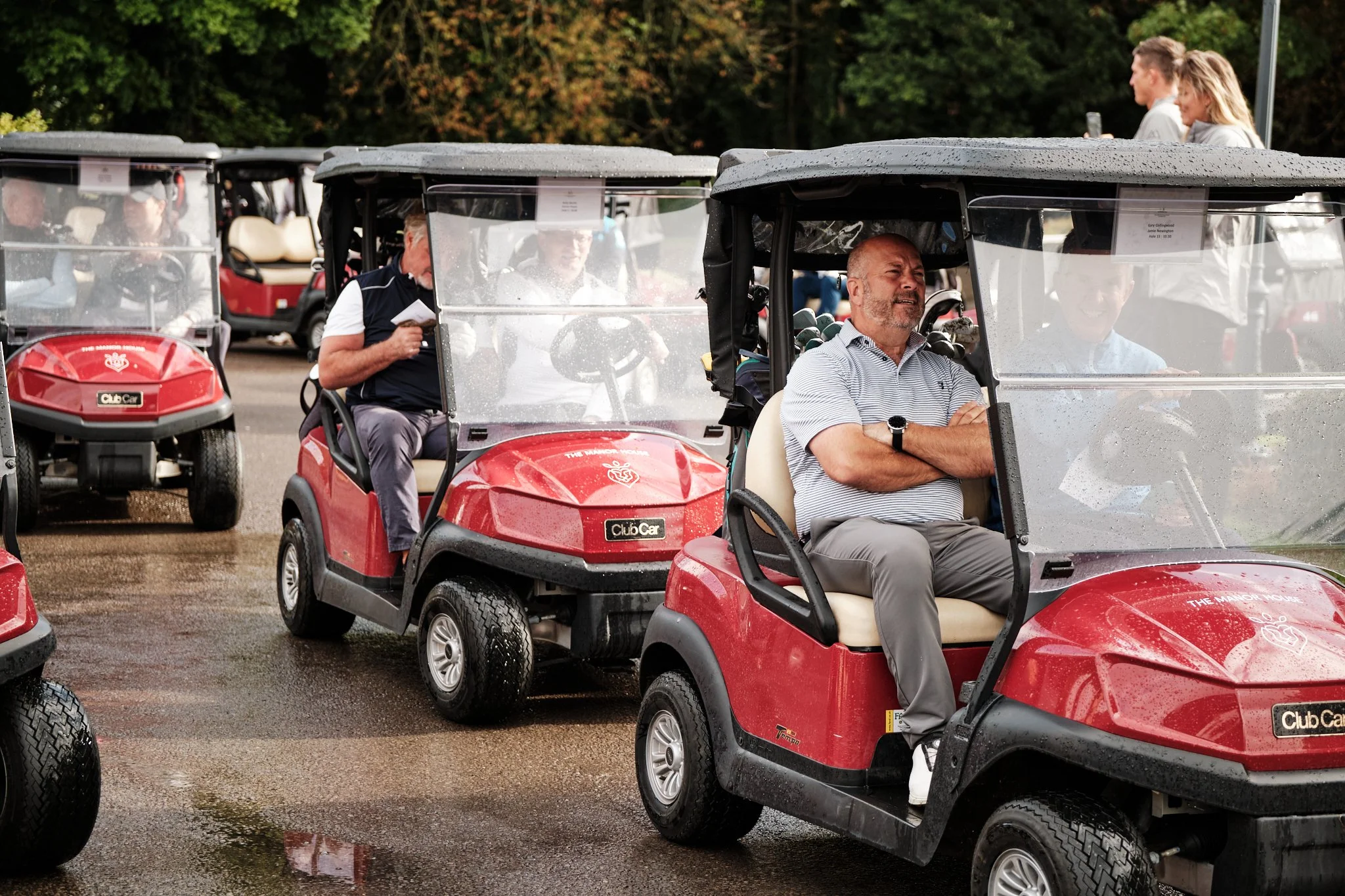 Several people riding in red golf carts on a wet golf course, with trees in the background. The front man appears to be a middle-aged man with a beard, sitting with arms crossed, looking up.