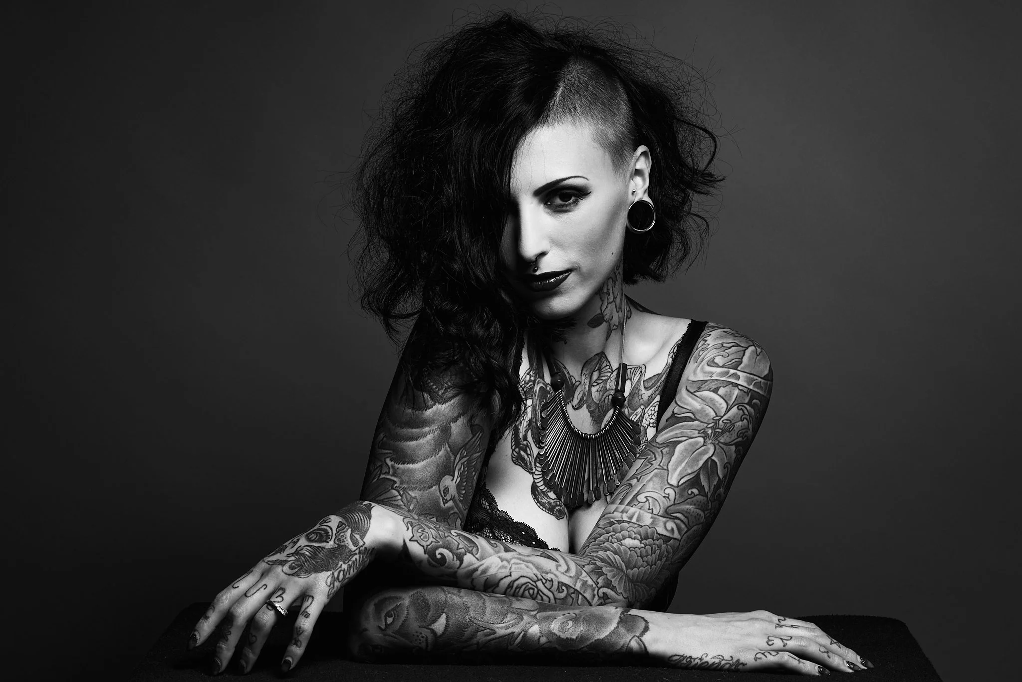 Black and white photo of a woman with tattoos, short hair shaved on one side, dark lipstick, and large earrings, sitting with her arms crossed on a surface, against a plain background.