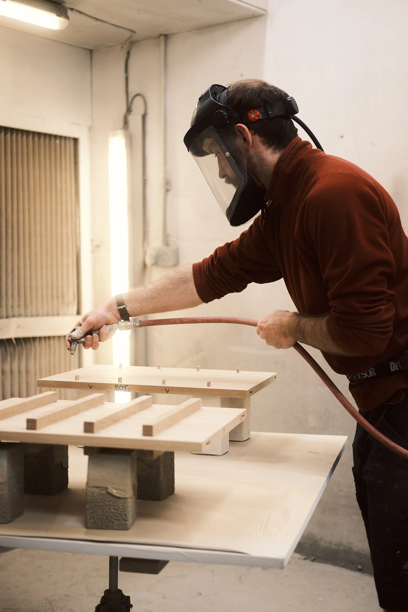 A man wearing a protective face shield and maroon shirt is spray painting wooden pieces on a table in a workshop.