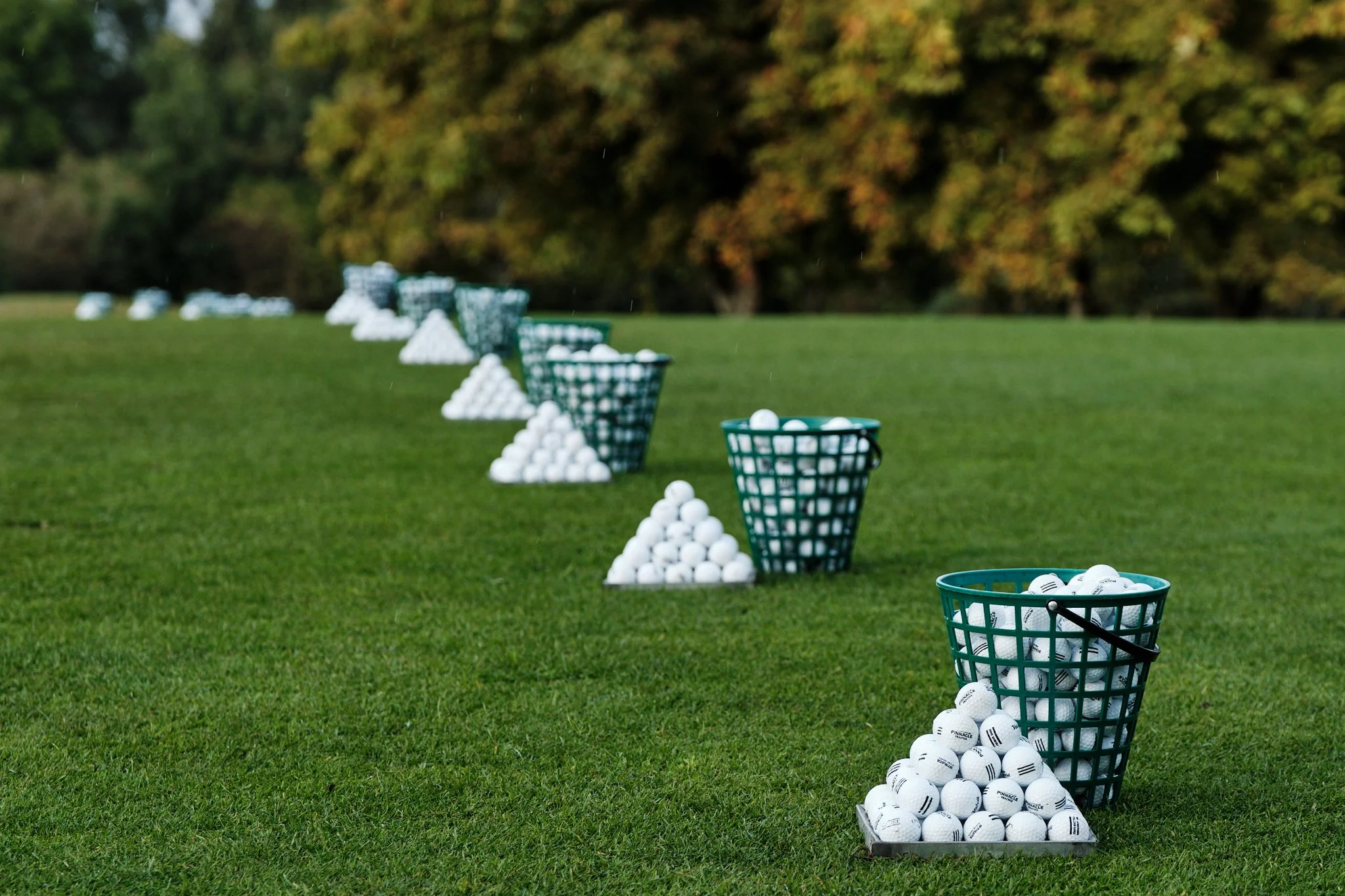 Multiple green baskets filled with white golf balls lined up on a golf course with a background of trees and grass.