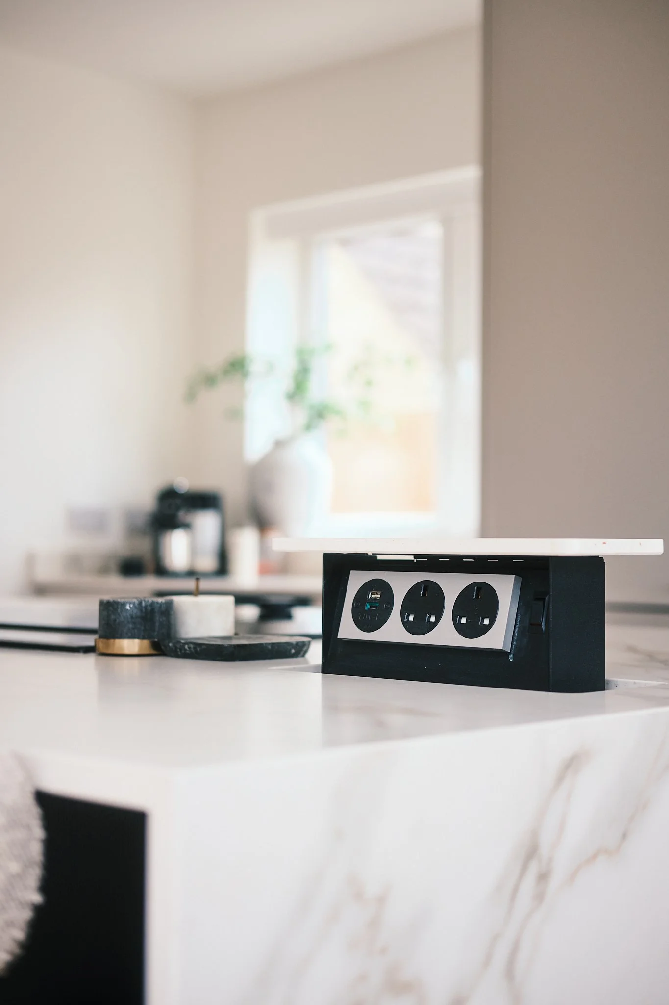 A kitchen countertop with a black power outlet with three sockets, a black and gold candle holder, and a black tray, with a background of a window, a potted plant, and coffee-making equipment.