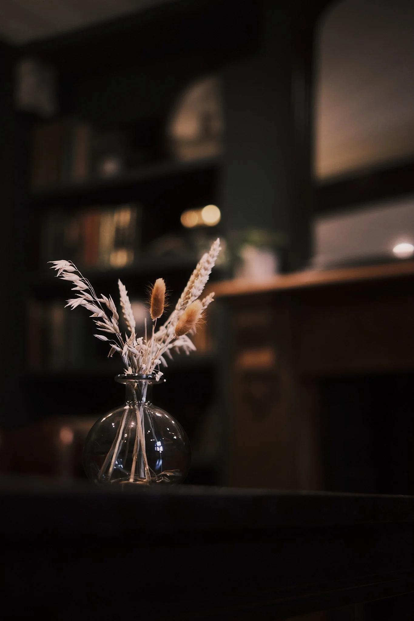 A clear glass vase with dried flowers including wheat stalks and fluffy grasses on a dark surface in a dimly lit room.