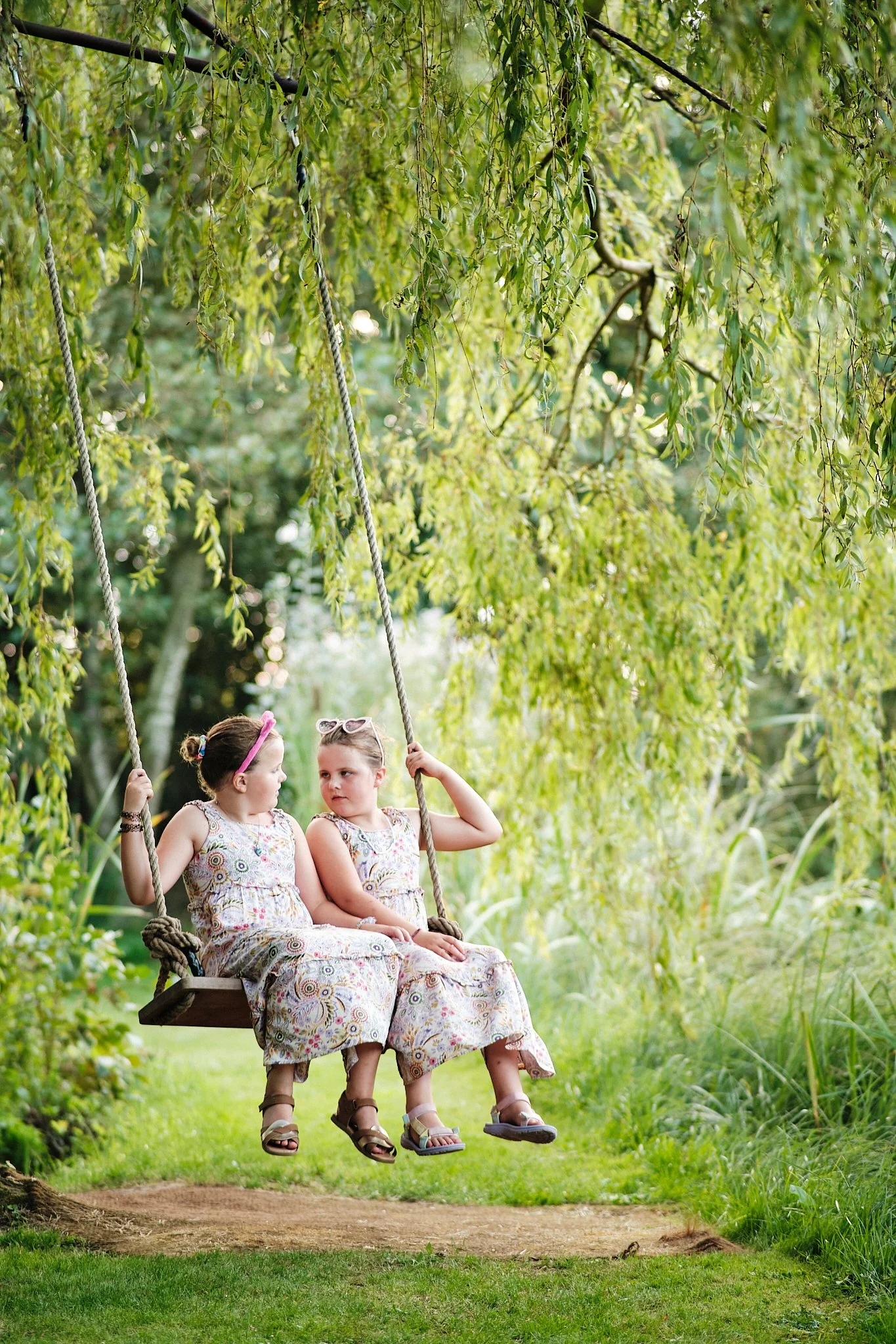 Two young girls sitting on a wooden swing hanging from a tree branch, surrounded by green foliage. The girls are wearing matching floral dresses and sandals, with one girl wearing sunglasses on her head. They appear to be having a conversation.