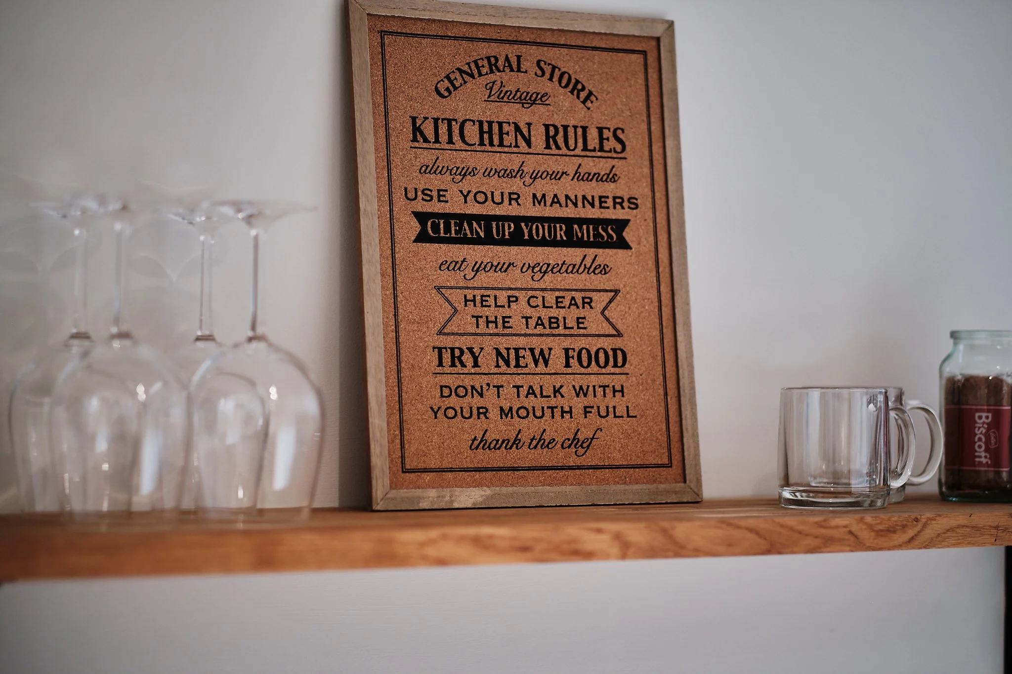 Kitchen rules sign on a wooden shelf, featuring rules such as wash hands, use manners, clean up mess, eat vegetables, clear table, try new food, not talk with full mouth, and thank the chef, with glasses and a mug beside the sign.