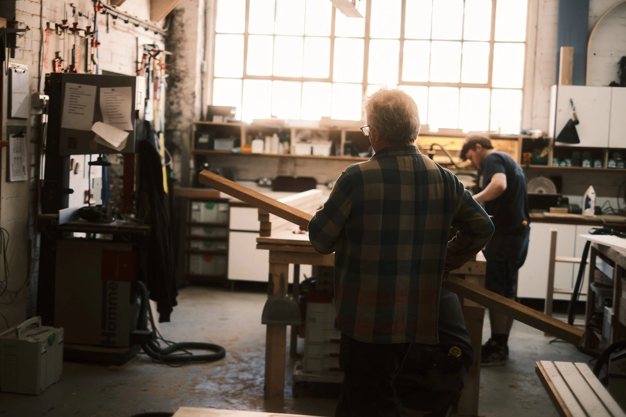 Two men working in a woodworking shop, one holding a piece of wood and the other working at a workbench, with large windows letting in natural light.