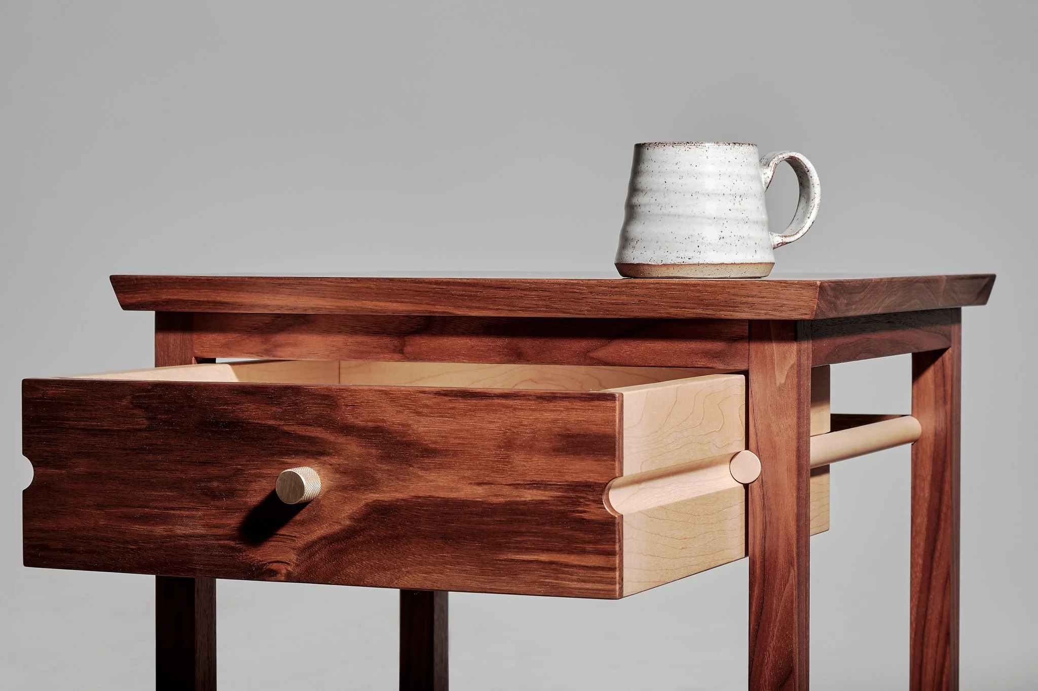 Close-up of a wooden table with an open drawer and a ceramic mug placed on top against a plain gray background.